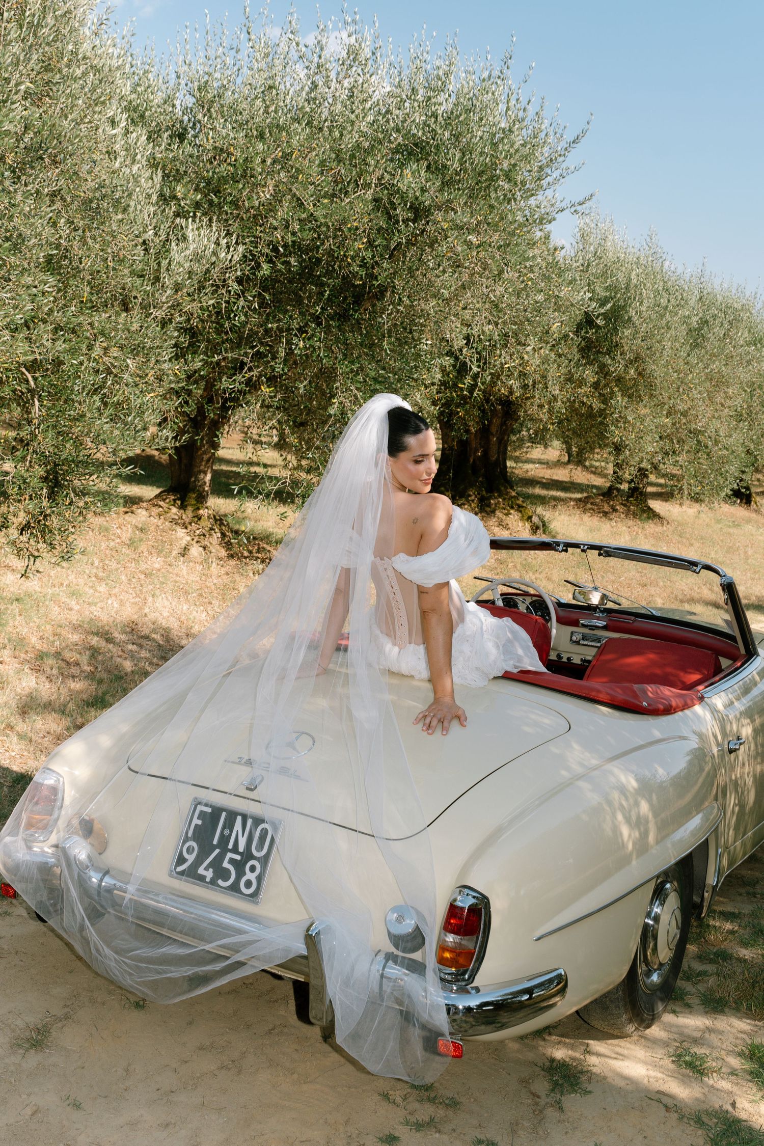 Bride in lace gown poses on vintage white convertible car surrounded by olive trees.