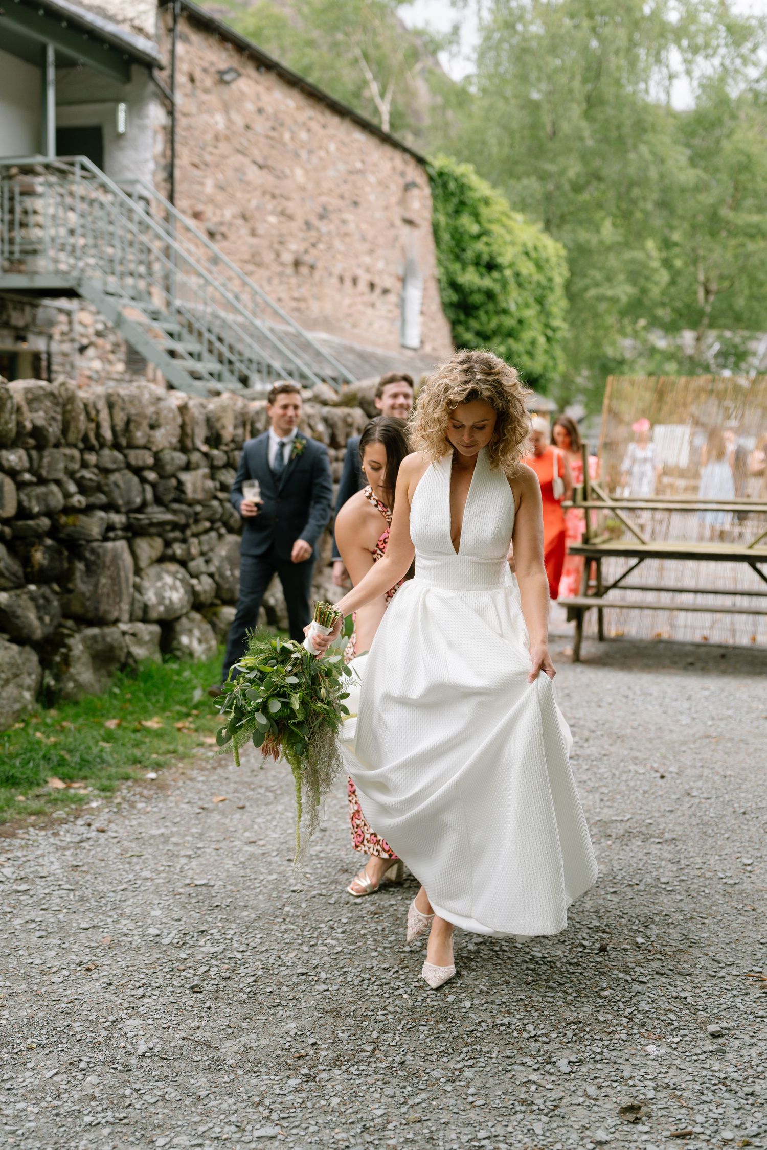 Wedding party walks along a gravel path beside a rustic stone building.