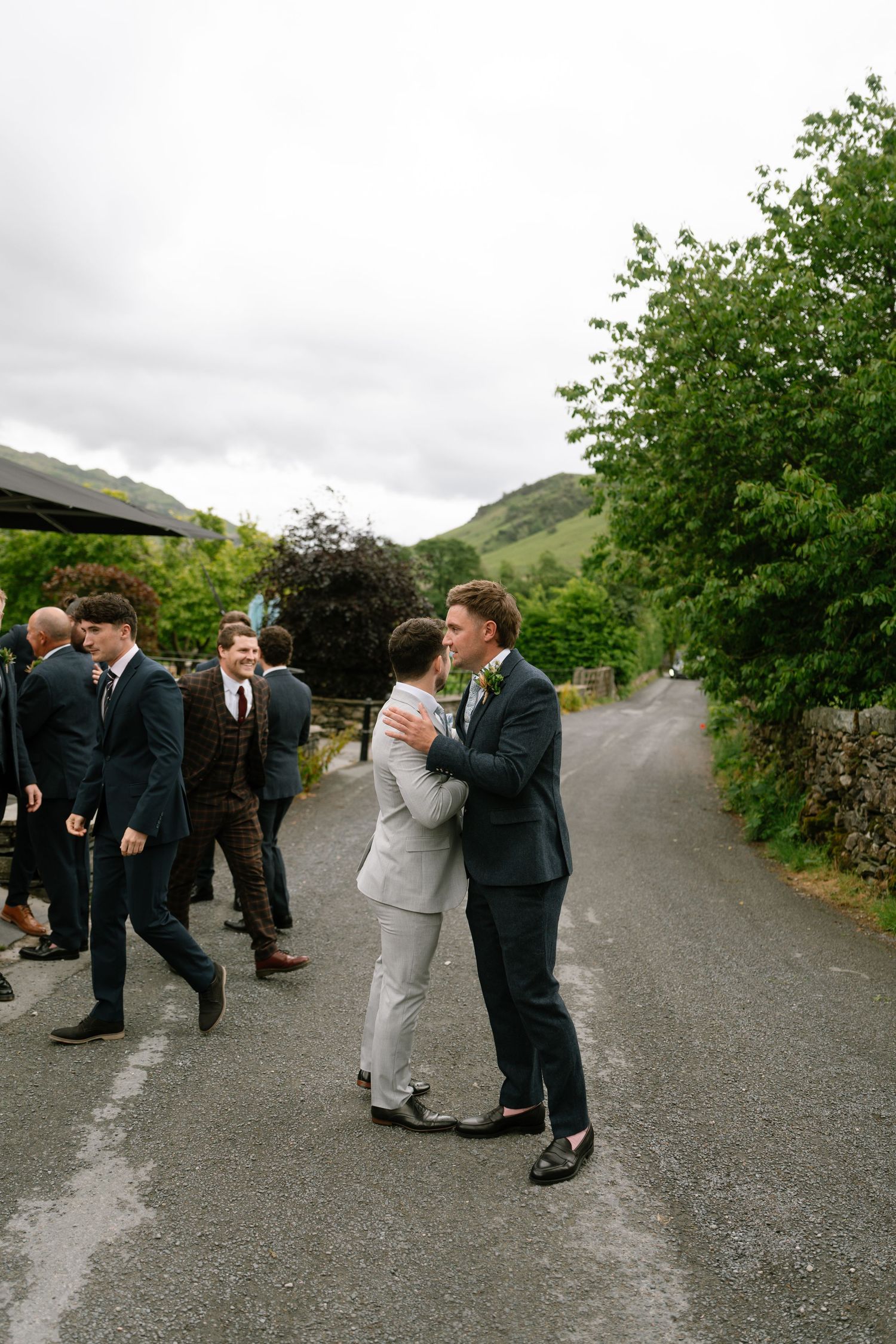 Wedding party in formal attire gathered on a rural road with green hills in the background.