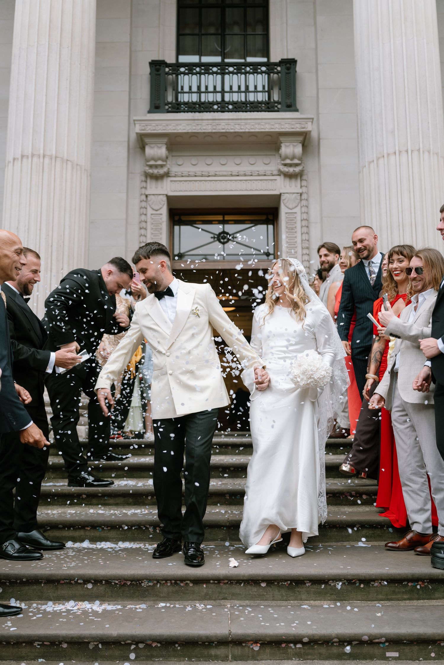 Wedding party and guests celebrating with confetti on grand steps of a classical building.