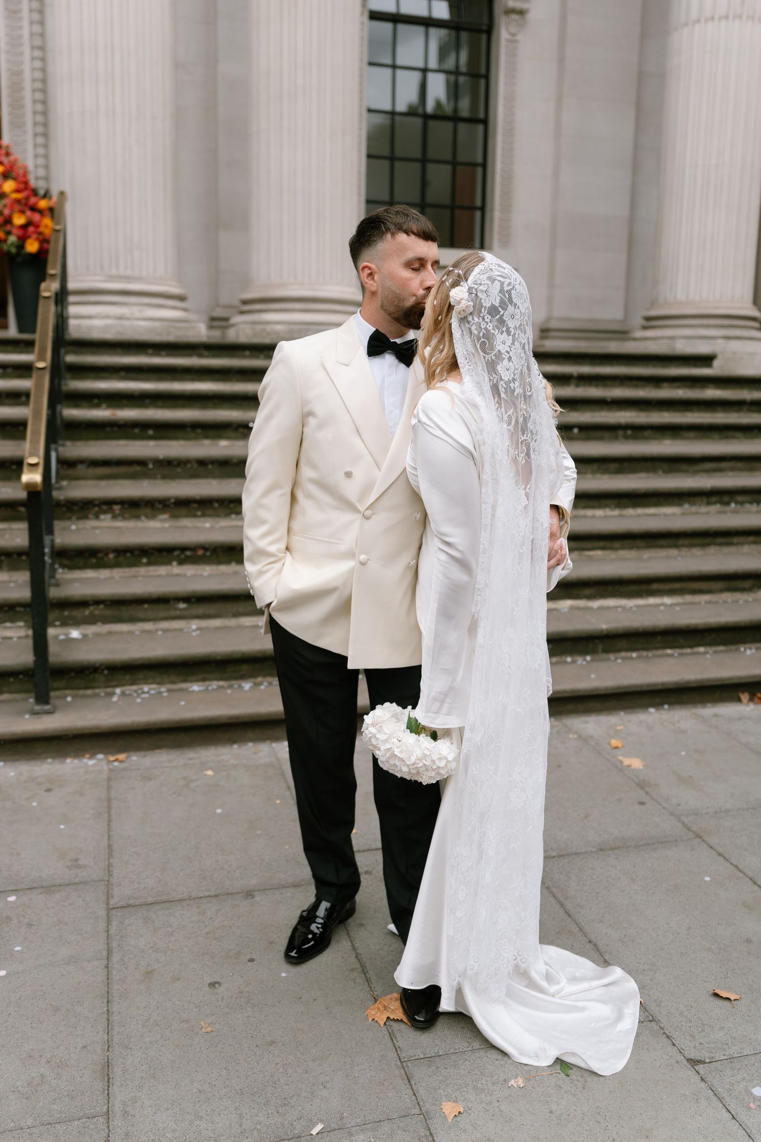 Couple in formal wedding attire walking outside a classical building with columns.