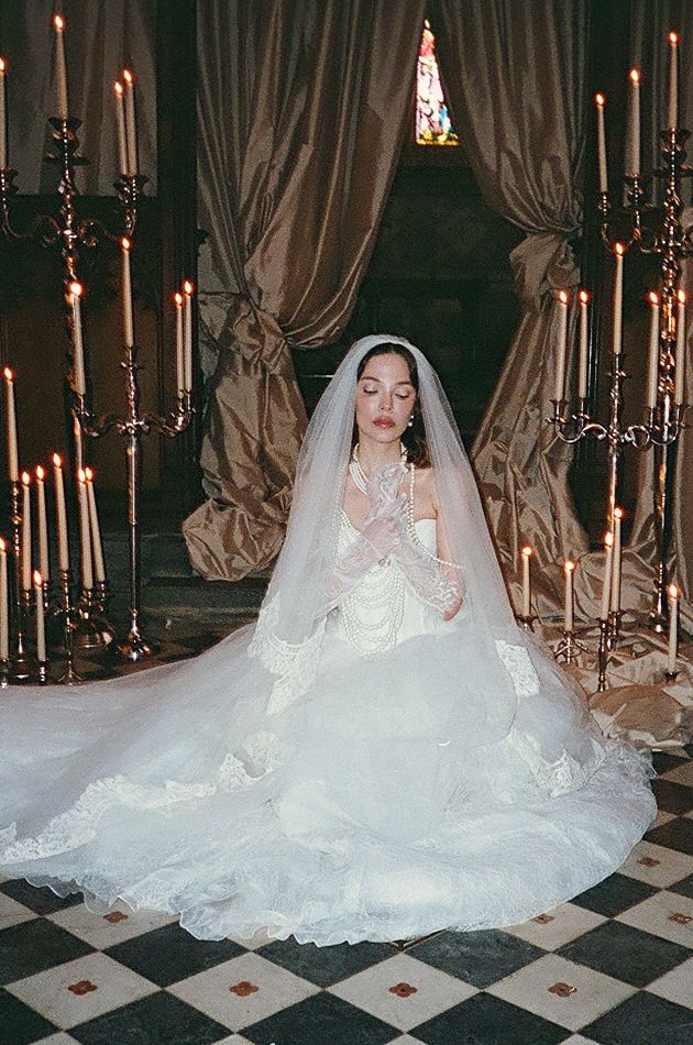 Bride in flowing white wedding gown and veil sits on checkered floor surrounded by glowing candles in elegant setting.