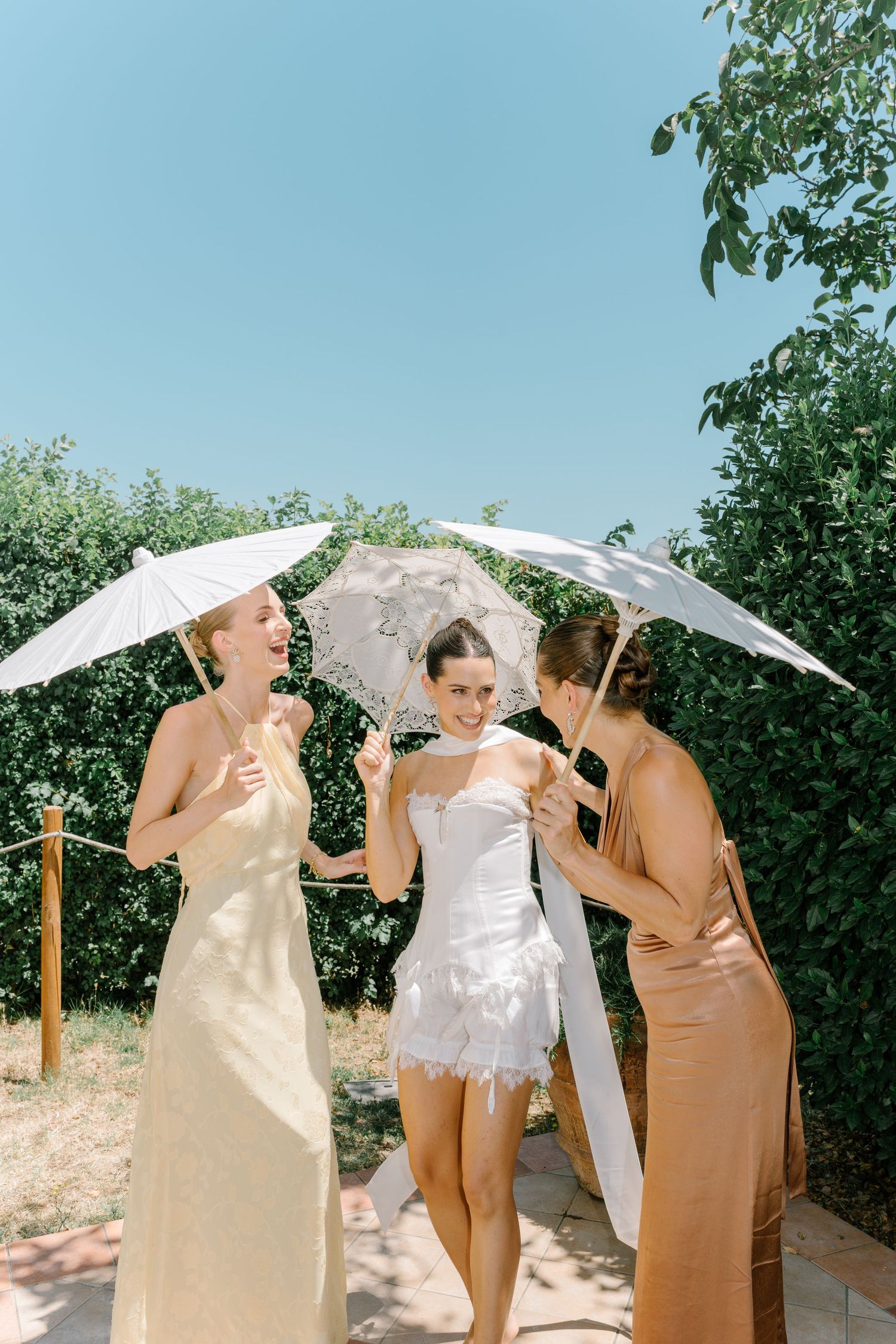 Bridesmaids in neutral-toned dresses pose with white parasols outdoors on a sunny day.