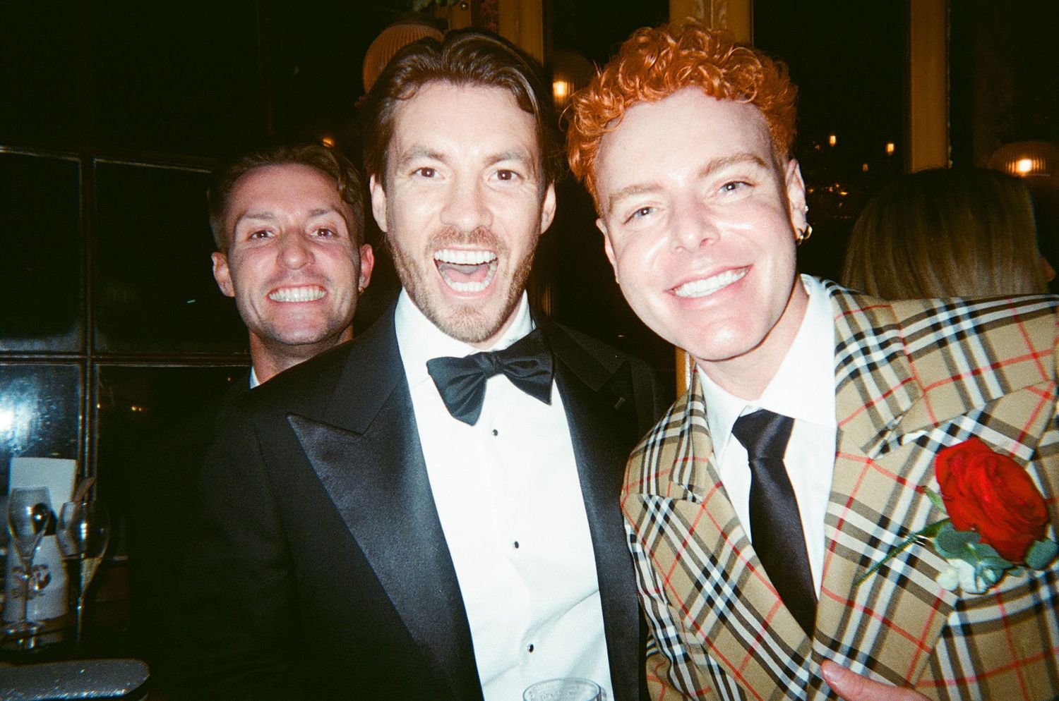 Three people in formal attire including tuxedos and a plaid jacket smile together at an evening event.