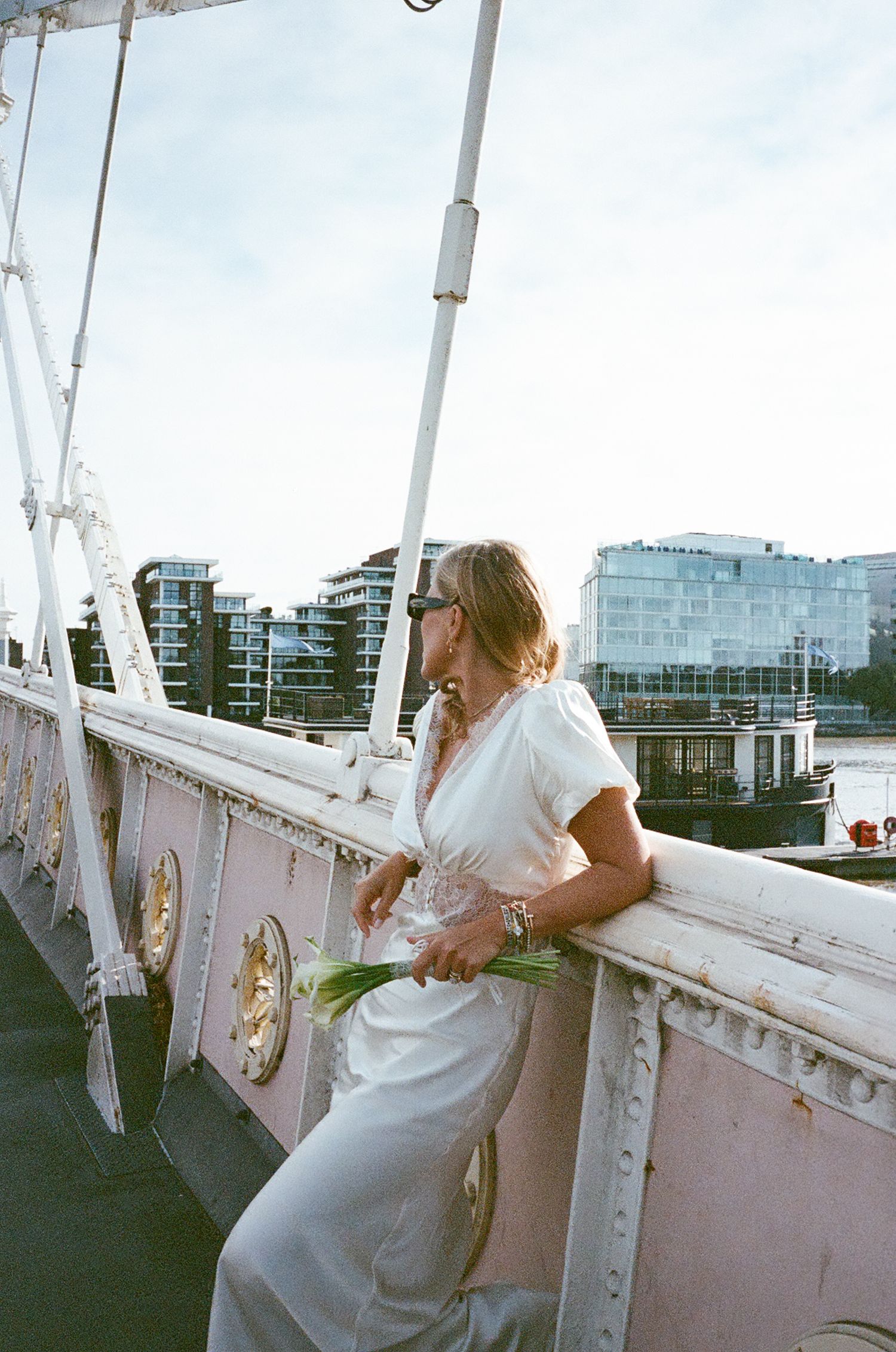A person in a white dress leans over the railing of a pink-painted bridge with modern buildings visible in the background.