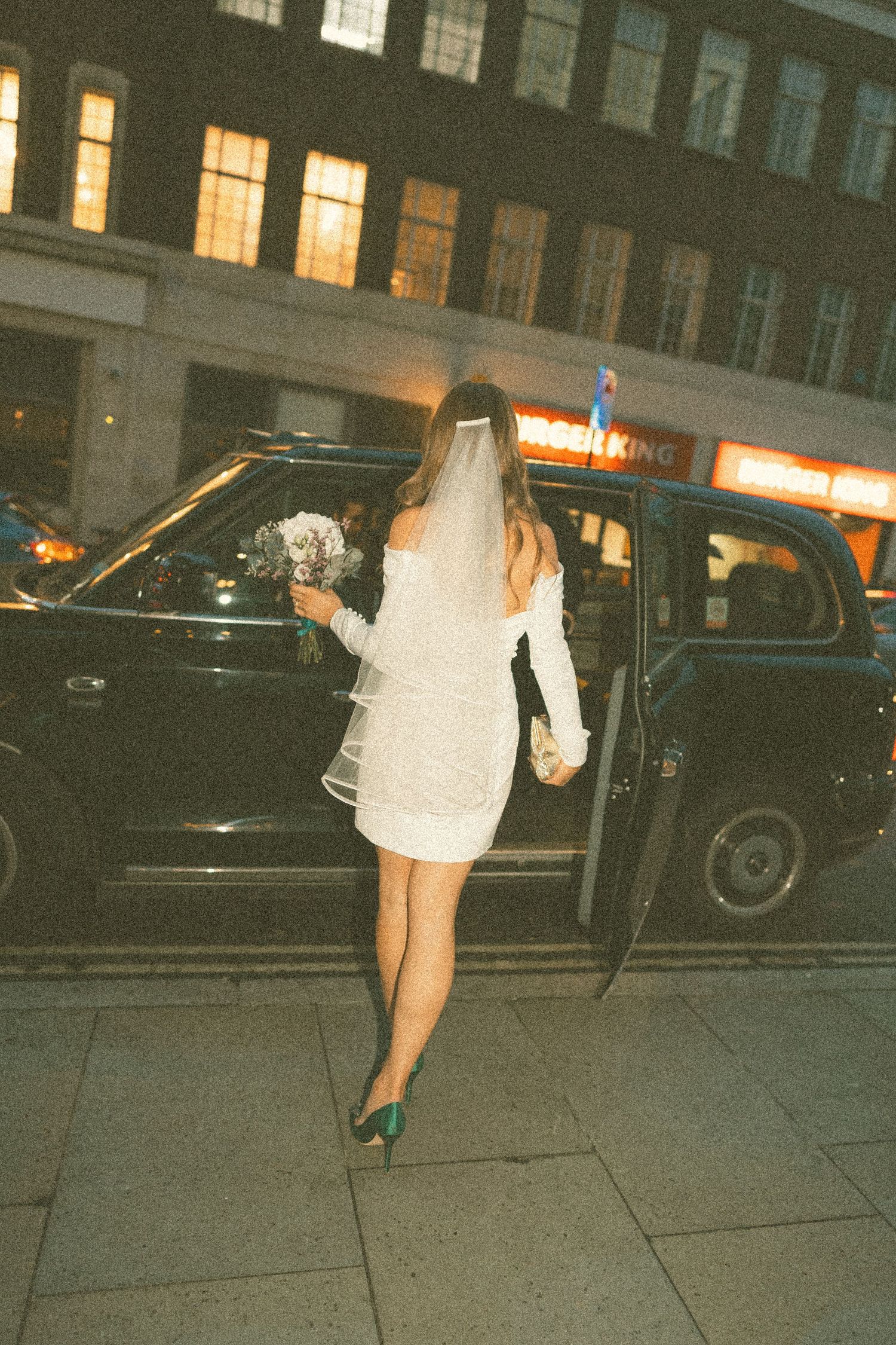A person in a short white dress and veil steps into a vintage black car on a city street at night.