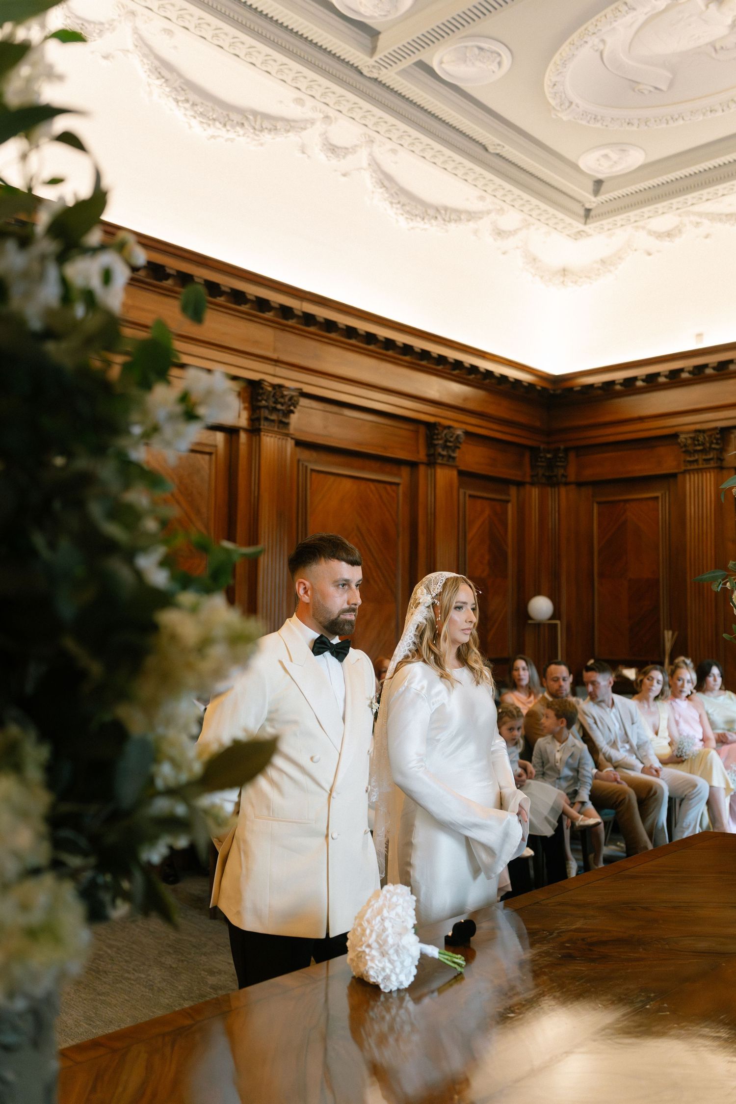 A wedding couple dances together in an elegant wood-paneled ballroom with ornate ceiling details.