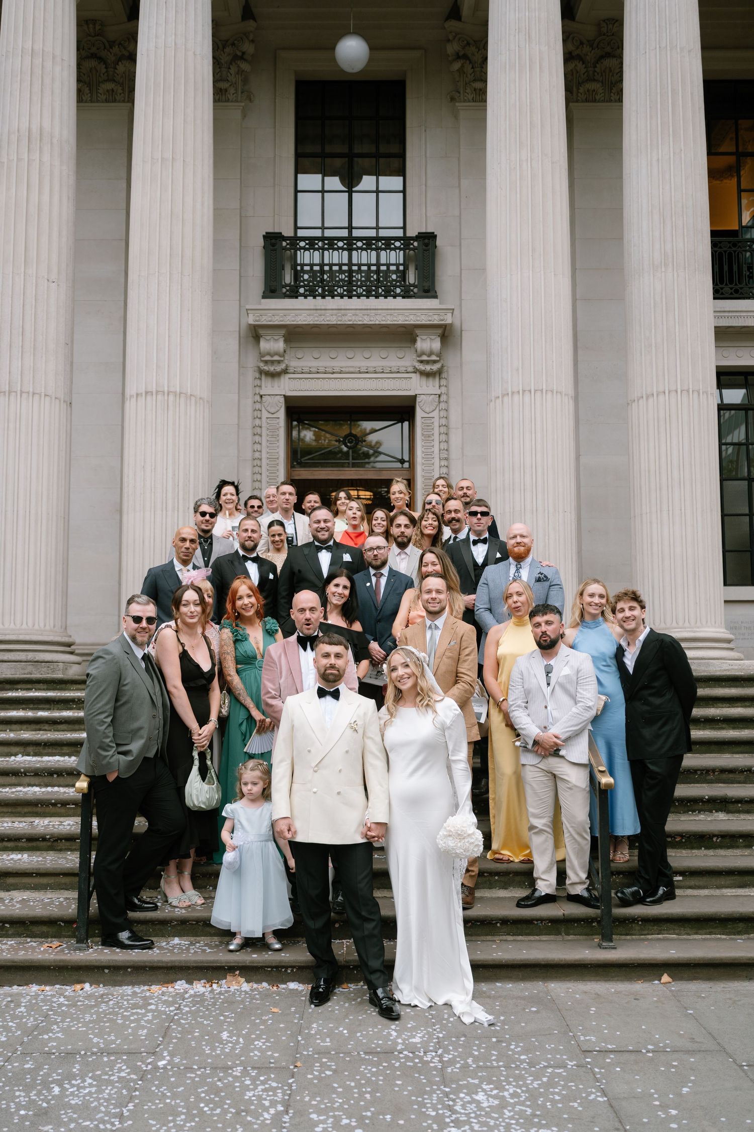 Wedding party gathers on grand stone steps of historic building with classical columns.