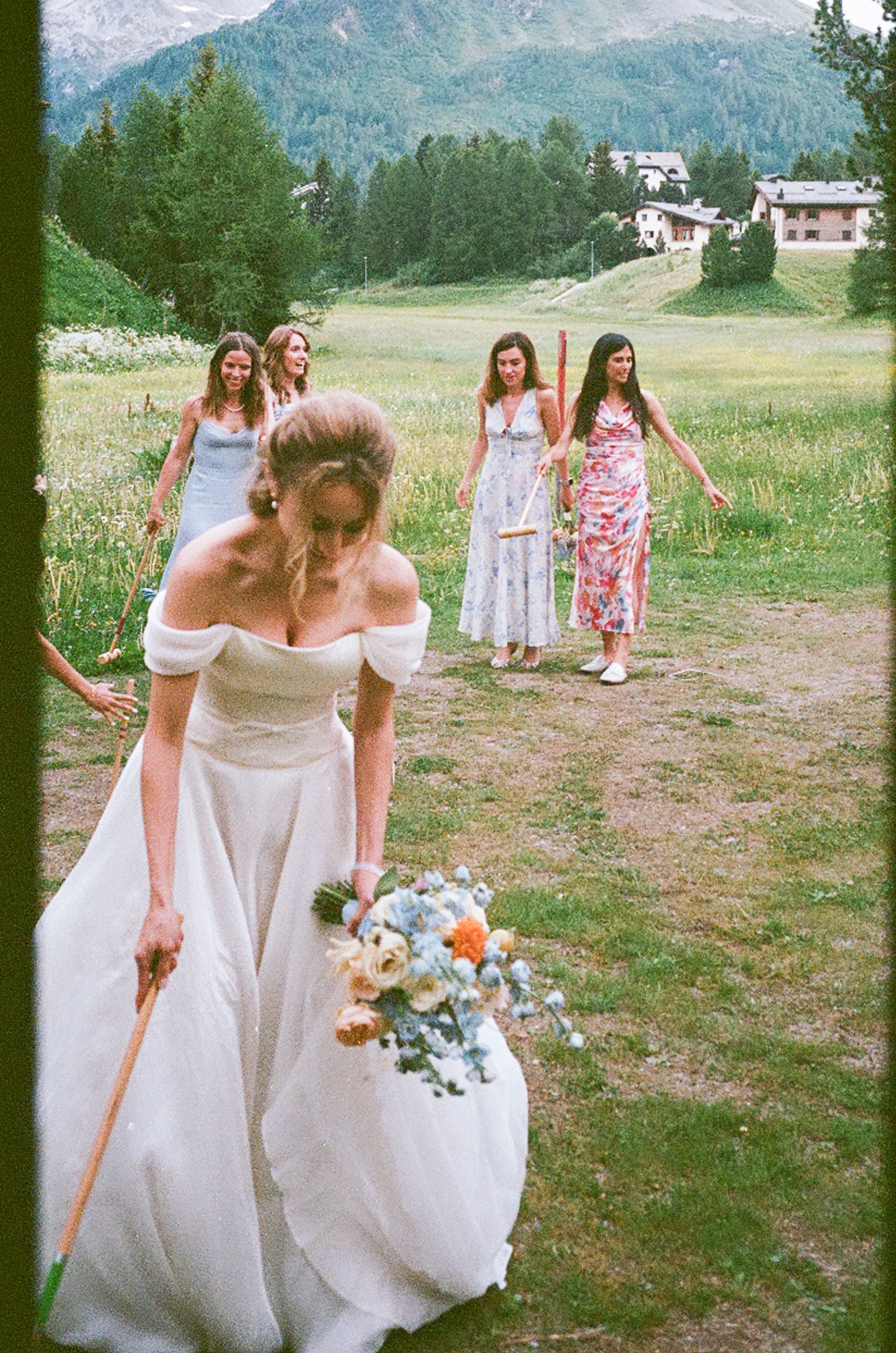 A wedding party walks across a grassy mountain meadow with evergreen forests and peaks in the background.