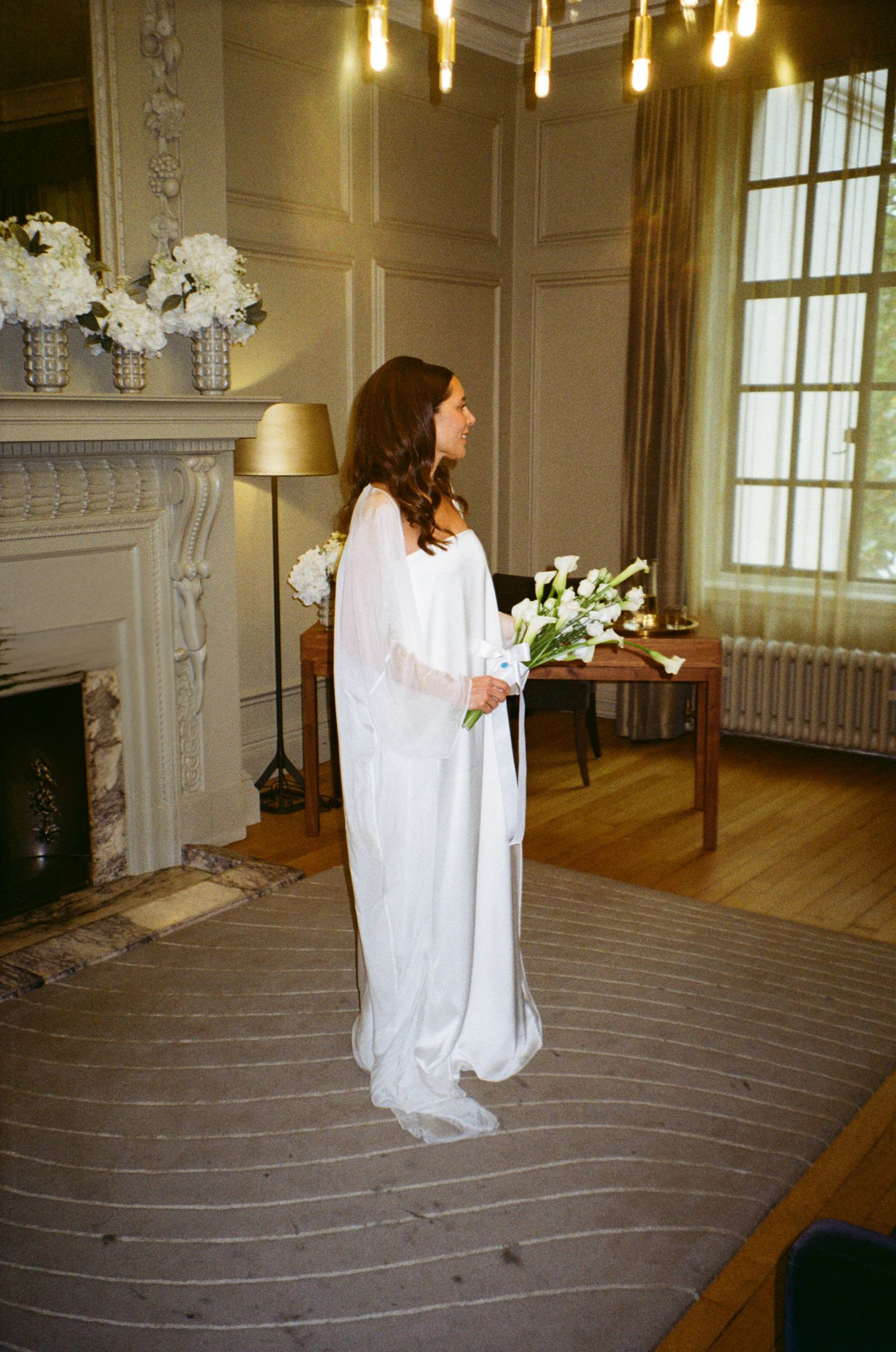 A flowing white wedding dress moves gracefully across a grand room with ornate walls and hanging light fixtures.