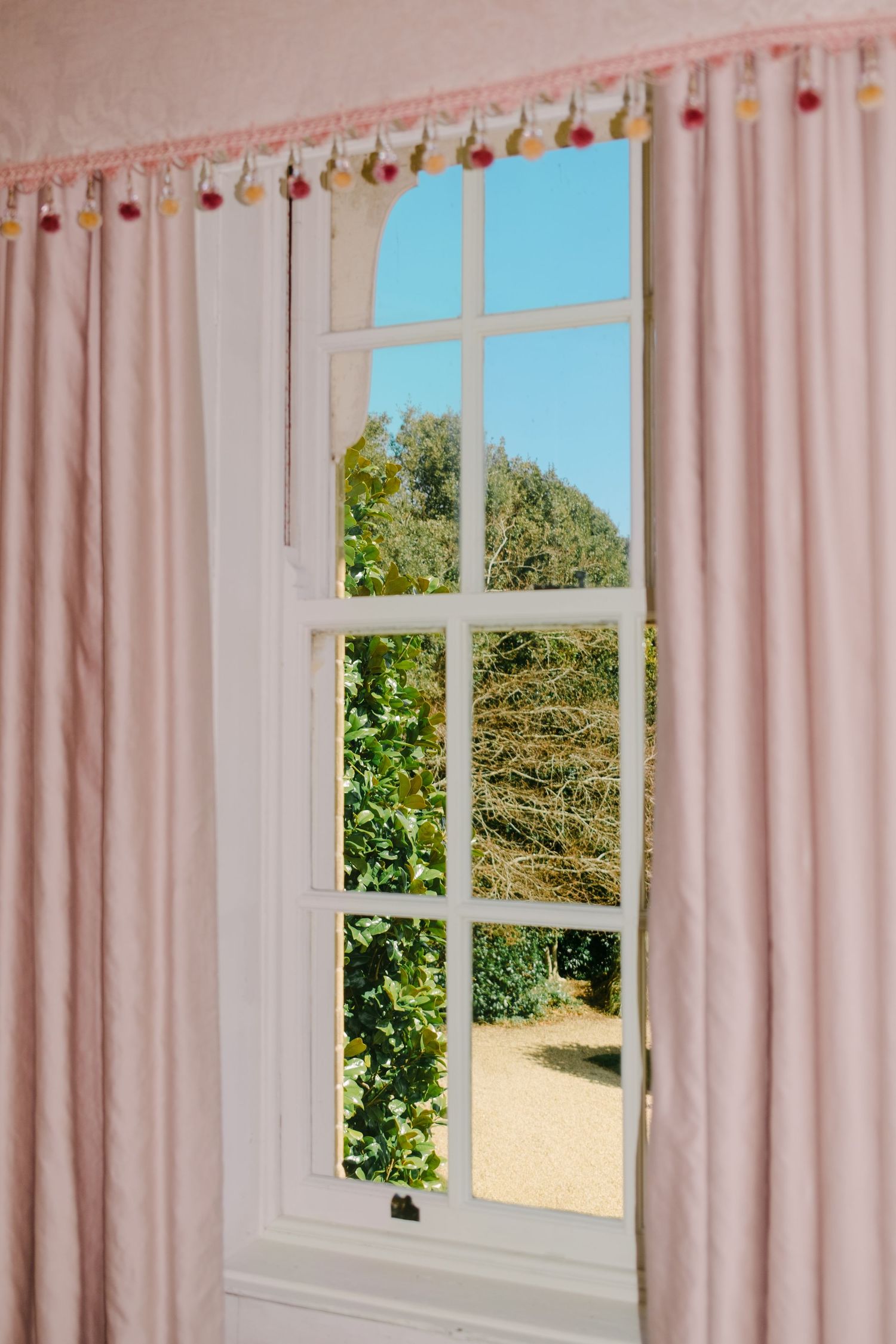 Pink curtains frame a bright window with string lights above, overlooking a sunny garden view with green trees.