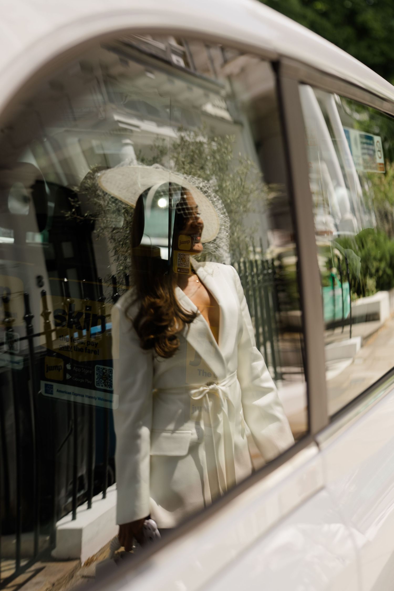 Elegant bride in white jacket and hat reflected in vintage car window on sunny day.