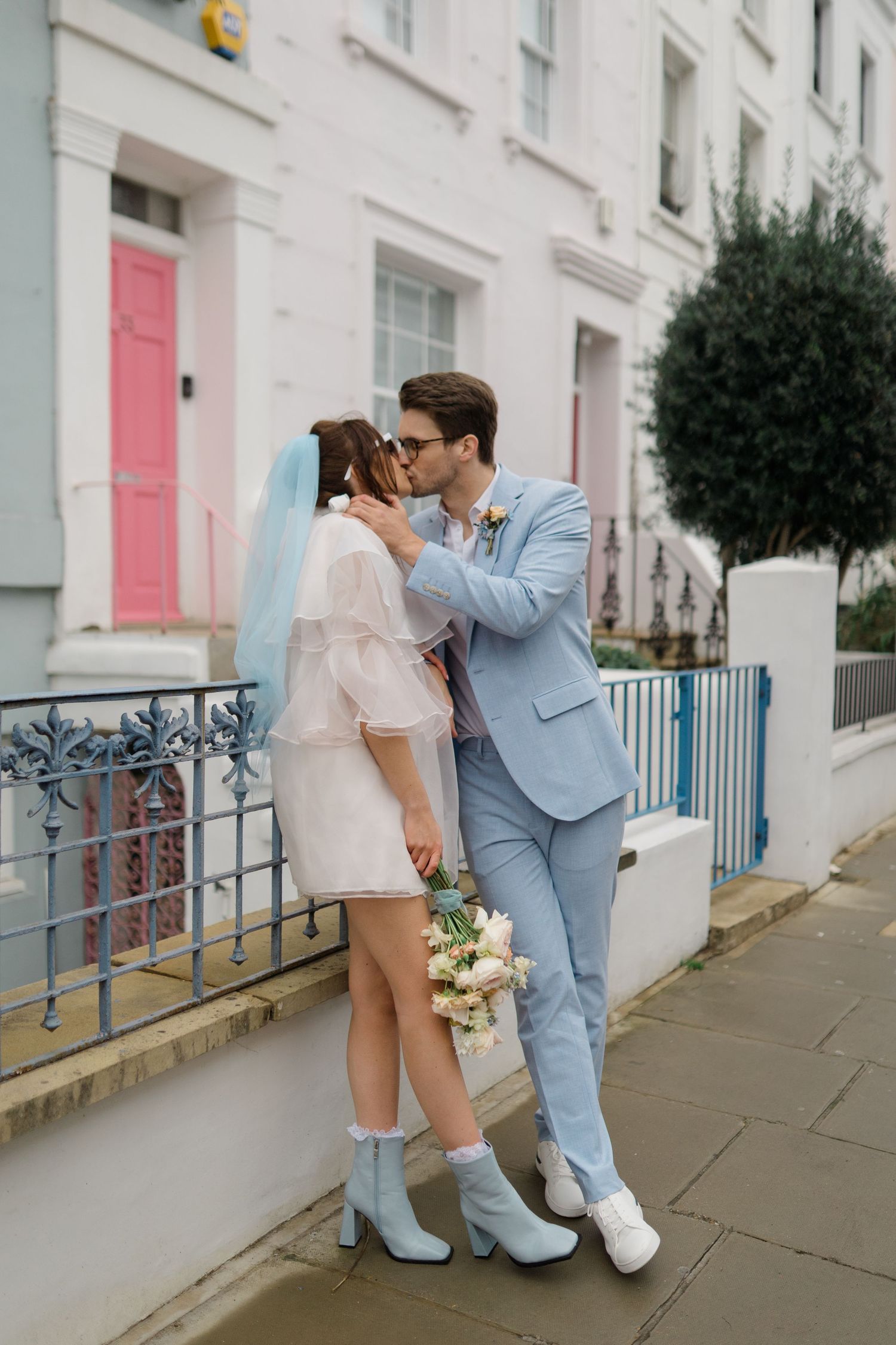 Bride and groom kissing on a colorful London street, she wears a short white dress with blue veil and boots.
