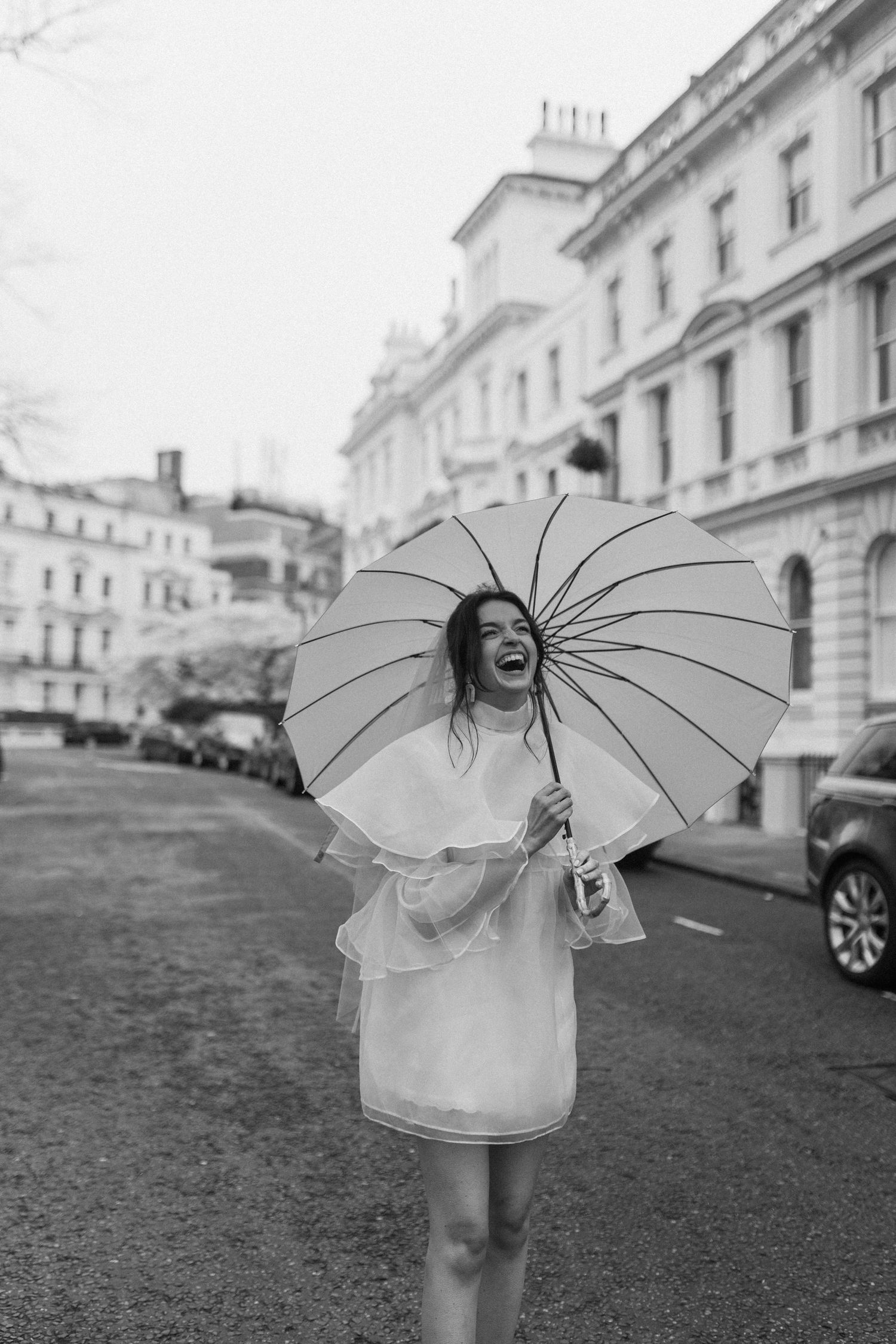 Black and white photo of a laughing woman holding a heart-shaped umbrella in a raincoat on a classic European street.