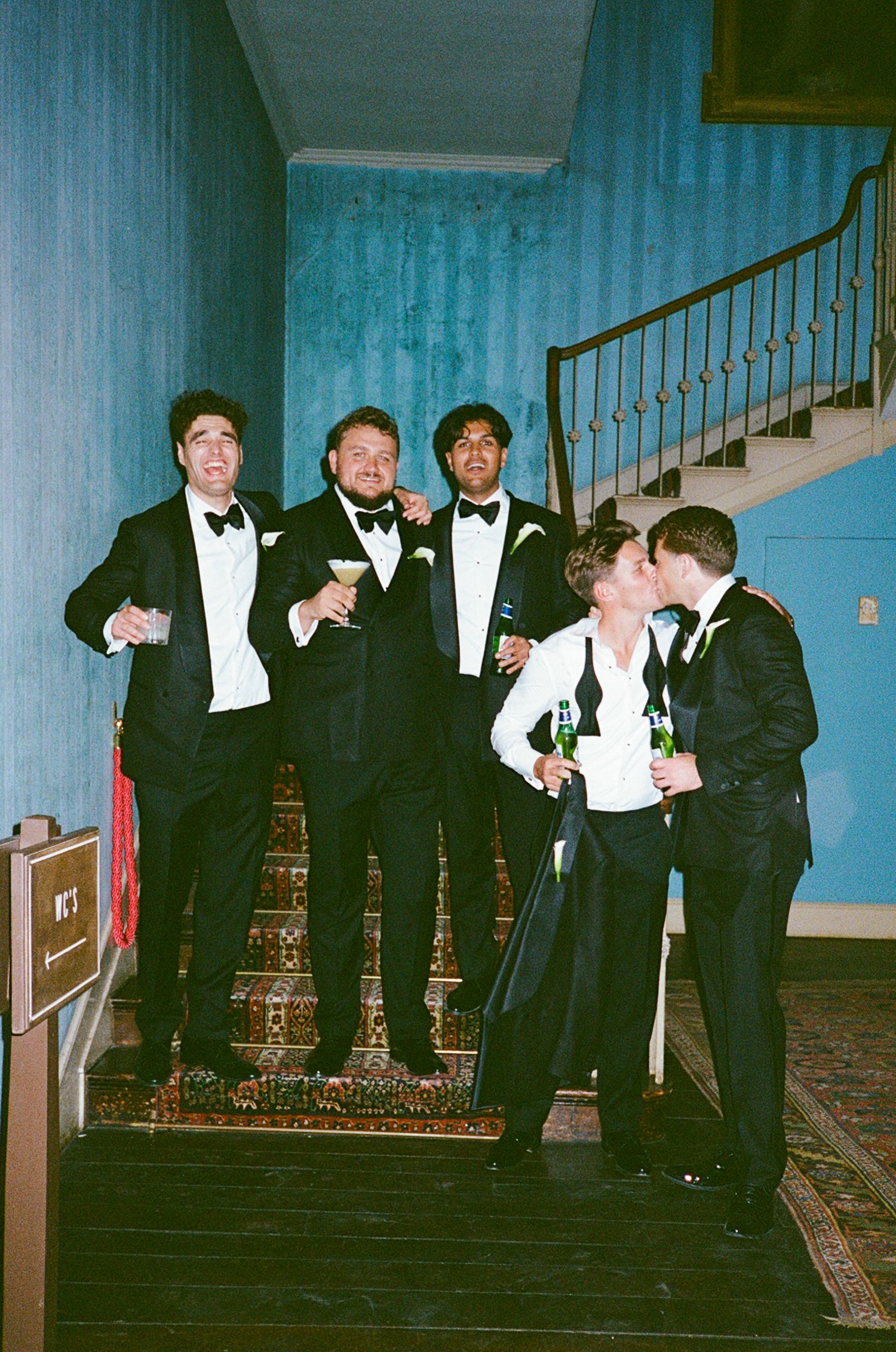 Five people in formal black tie attire pose on a staircase, holding drinks at an elegant evening event.
