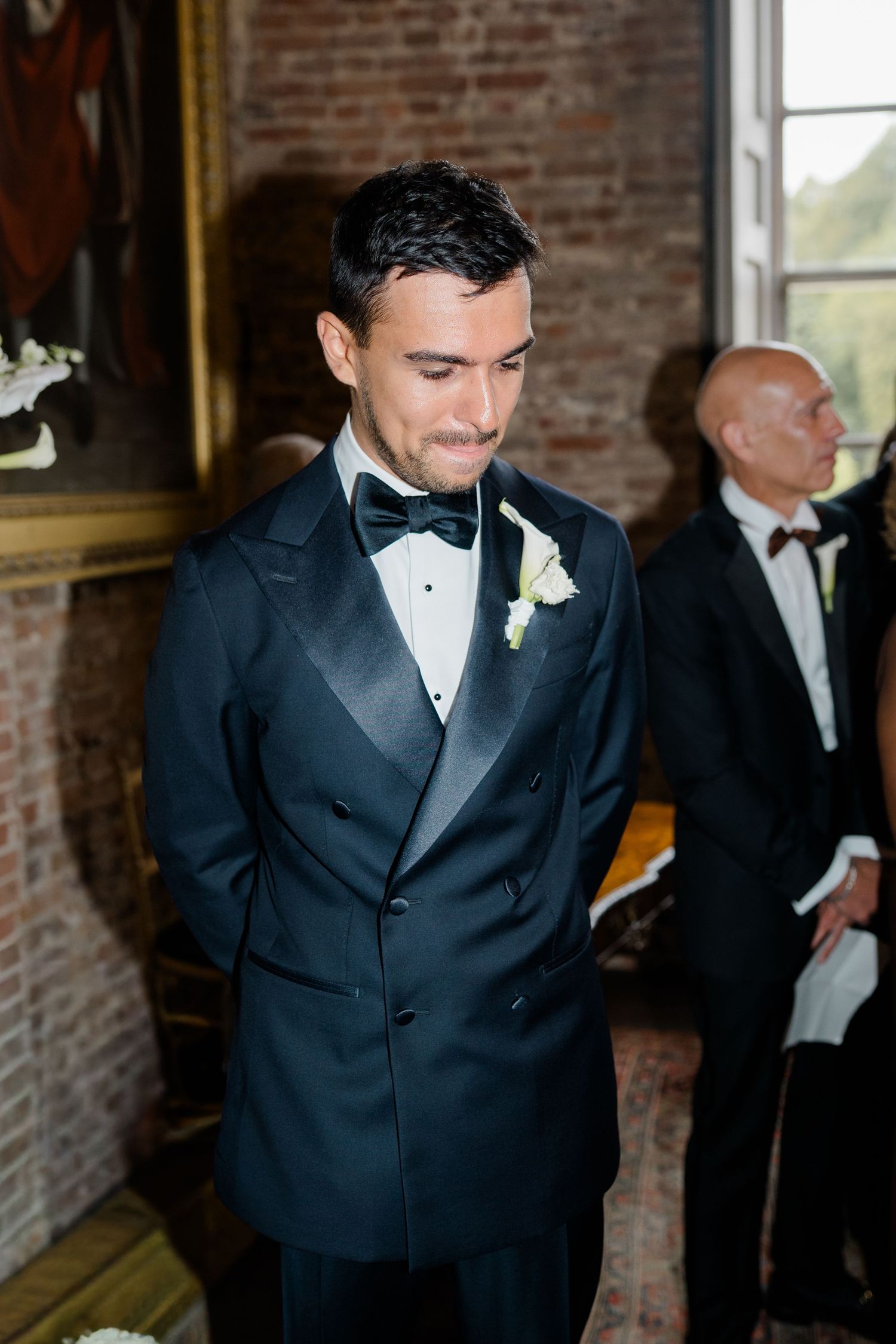 Groom in double-breasted black tuxedo with white boutonniere stands at wedding ceremony altar.