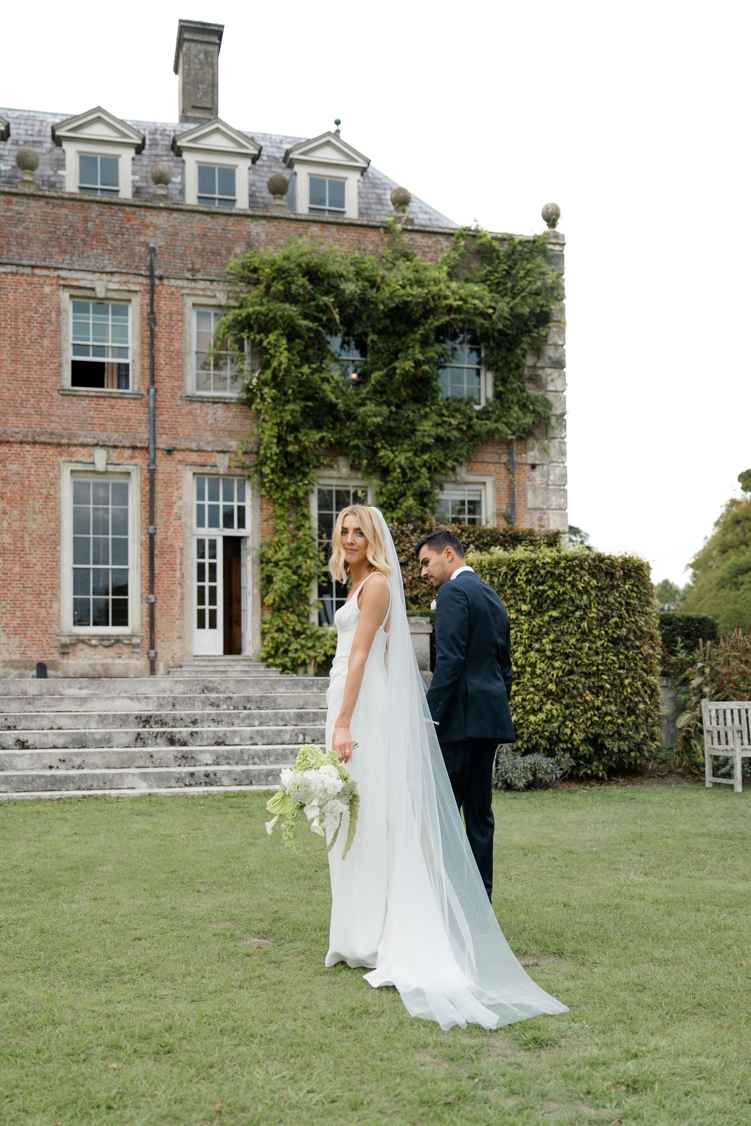 Bride in flowing veil and groom stand on lawn before ivy-covered Georgian country house venue.