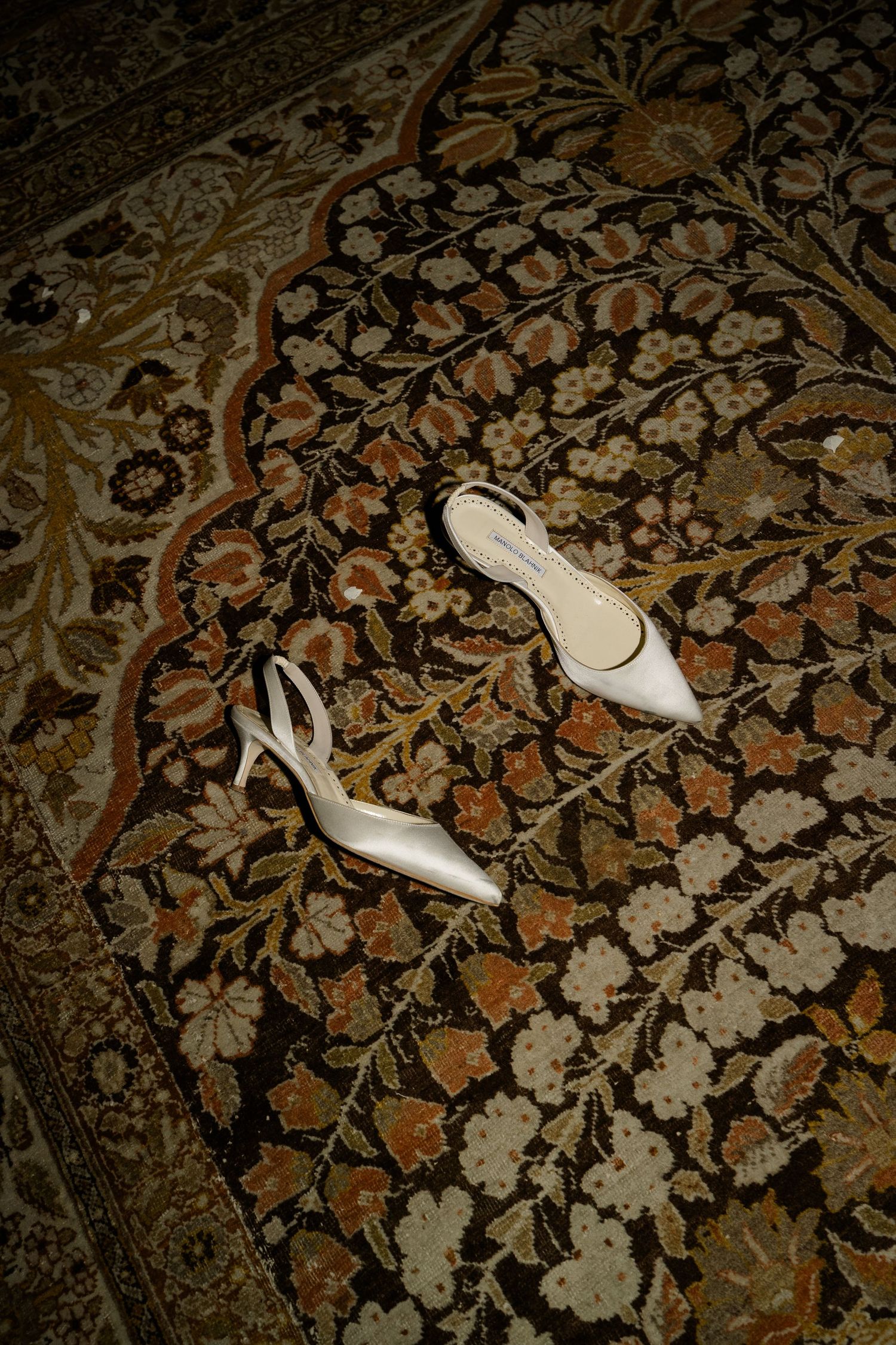 White satin slingback heels placed on an ornate floral patterned rug with rich earth tones.