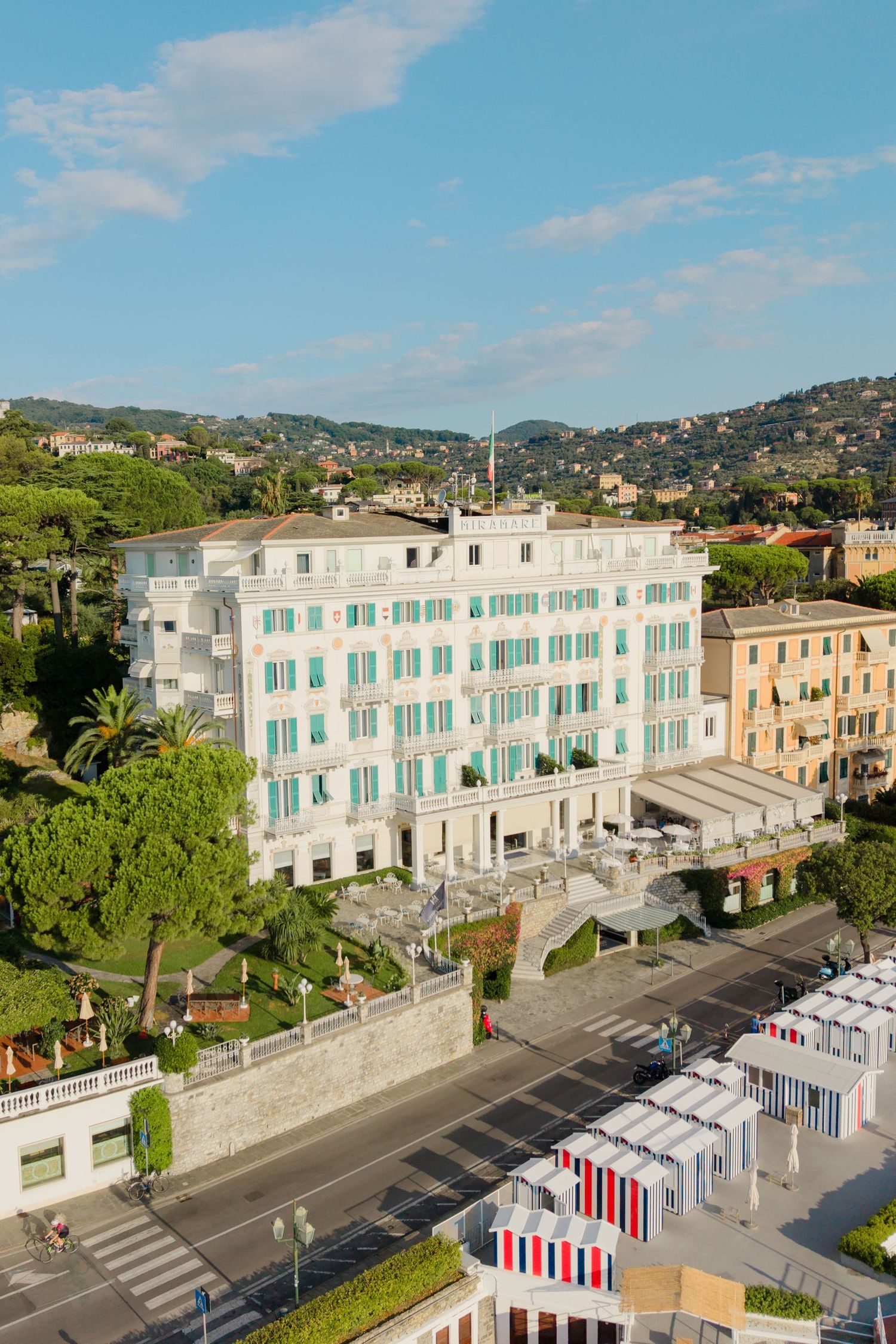 Elegant white hotel facade with turquoise shutters overlooking a coastal Italian town with lush green hills.