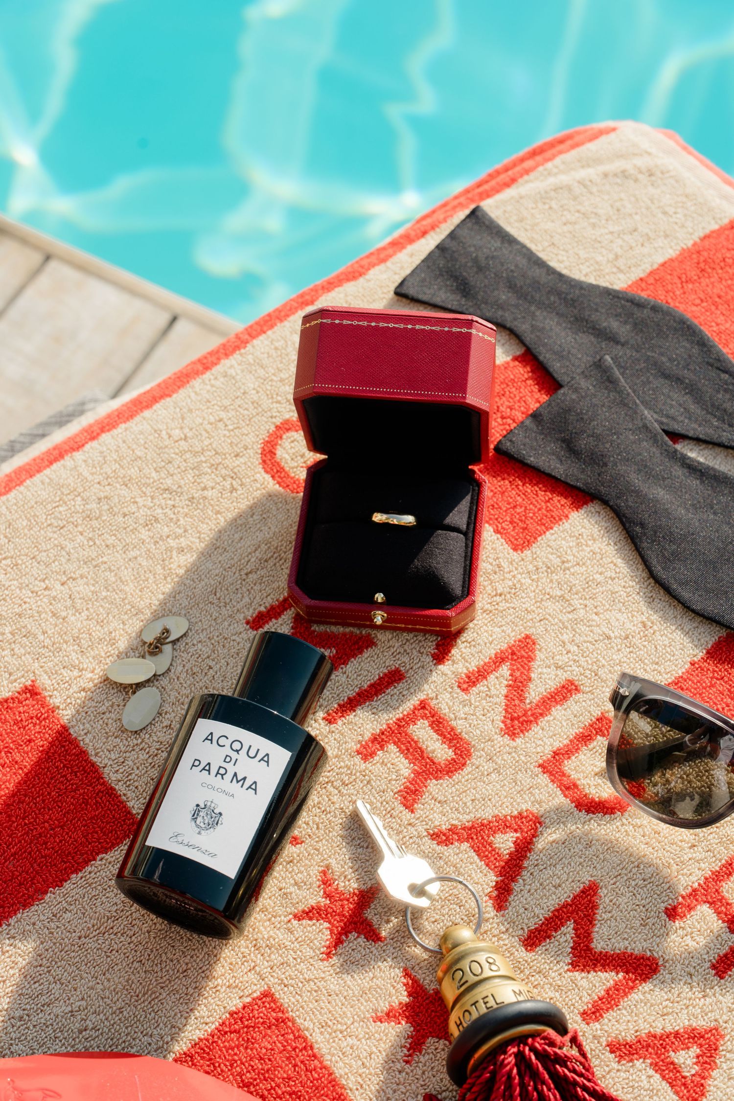 Acqua di Parma cologne, Cartier ring box, sunglasses, and champagne bottle on a striped beach towel by a pool.