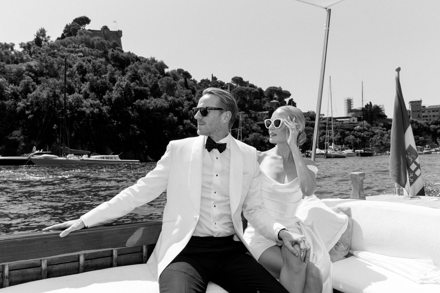 Bride and groom in wedding attire sit on luxury boat with scenic Italian coastline backdrop.