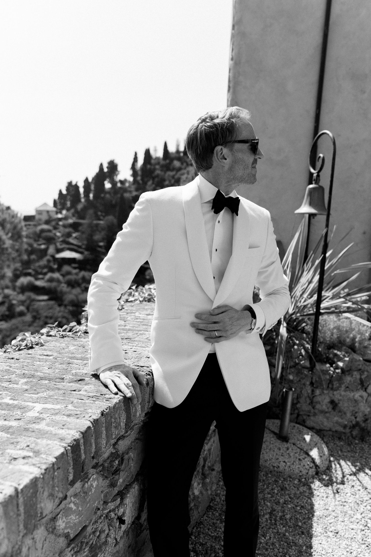 Sophisticated man in white tuxedo jacket and black bow tie posing outdoors in black and white photo.
