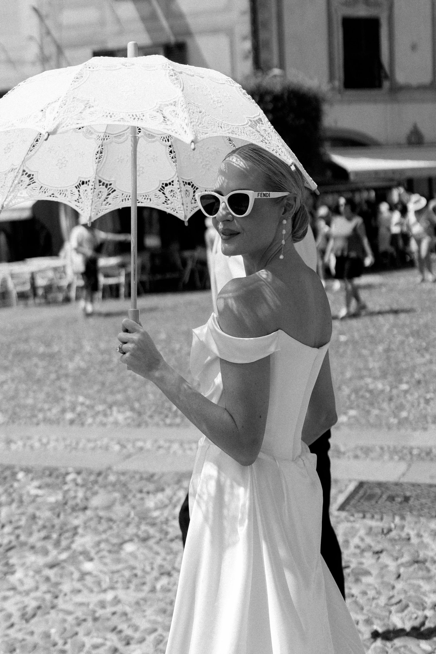 Elegant bride in white off-shoulder dress wearing cat-eye sunglasses holding lace parasol outdoors.