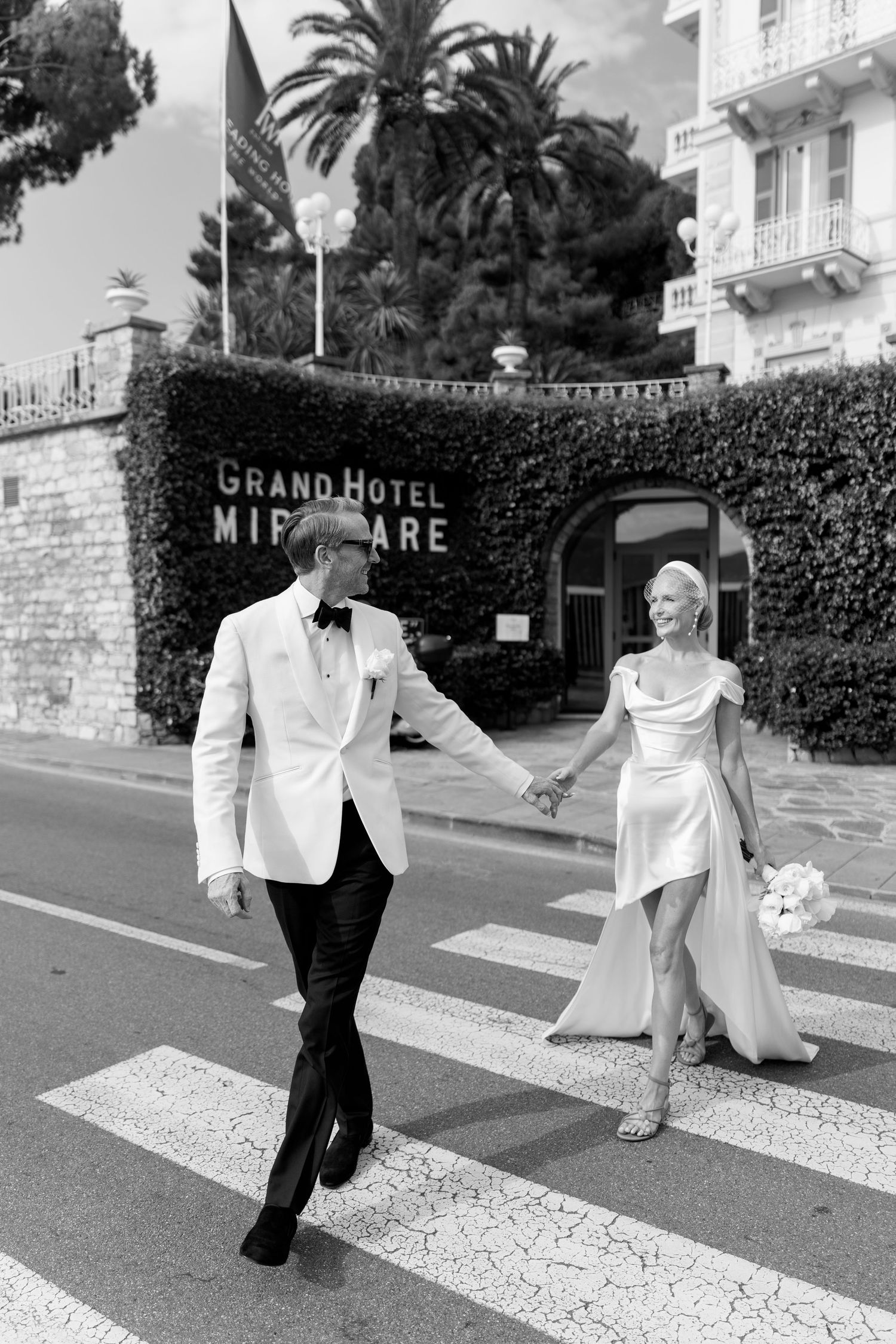 Bride and groom cross street holding hands outside Grand Hotel Miramare in black and white.