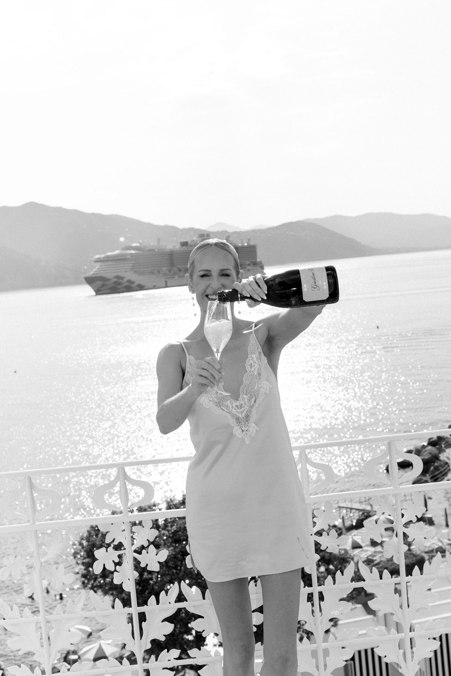 Glamorous blonde woman pouring champagne on a balcony with a cruise ship and sparkling sea behind her.