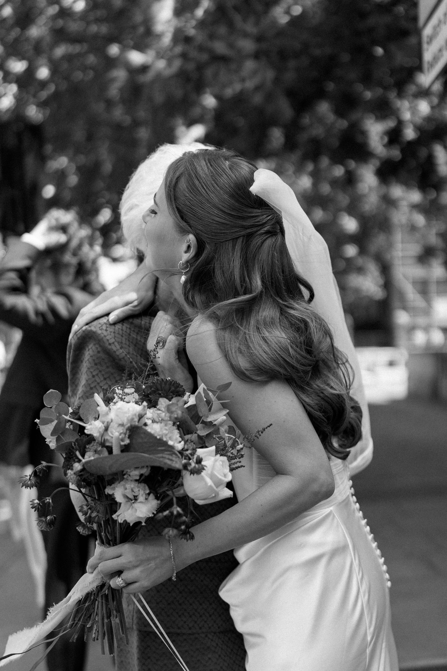 Bride in white gown and veil embraces guest outdoors, holding a beautiful floral bouquet in black and white photo.