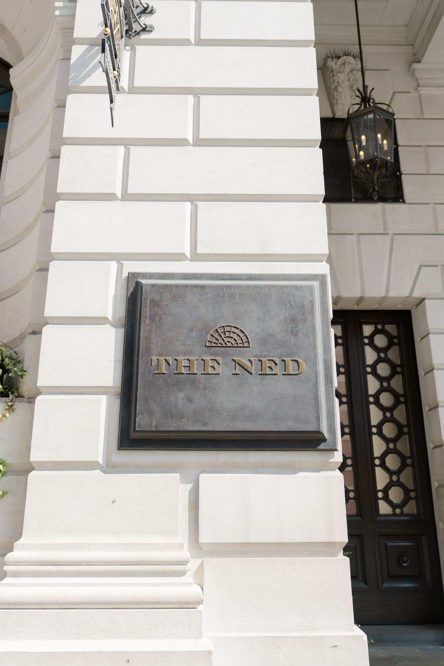 The Ned hotel entrance featuring an elegant bronze sign on white stone facade with ornate black lantern.