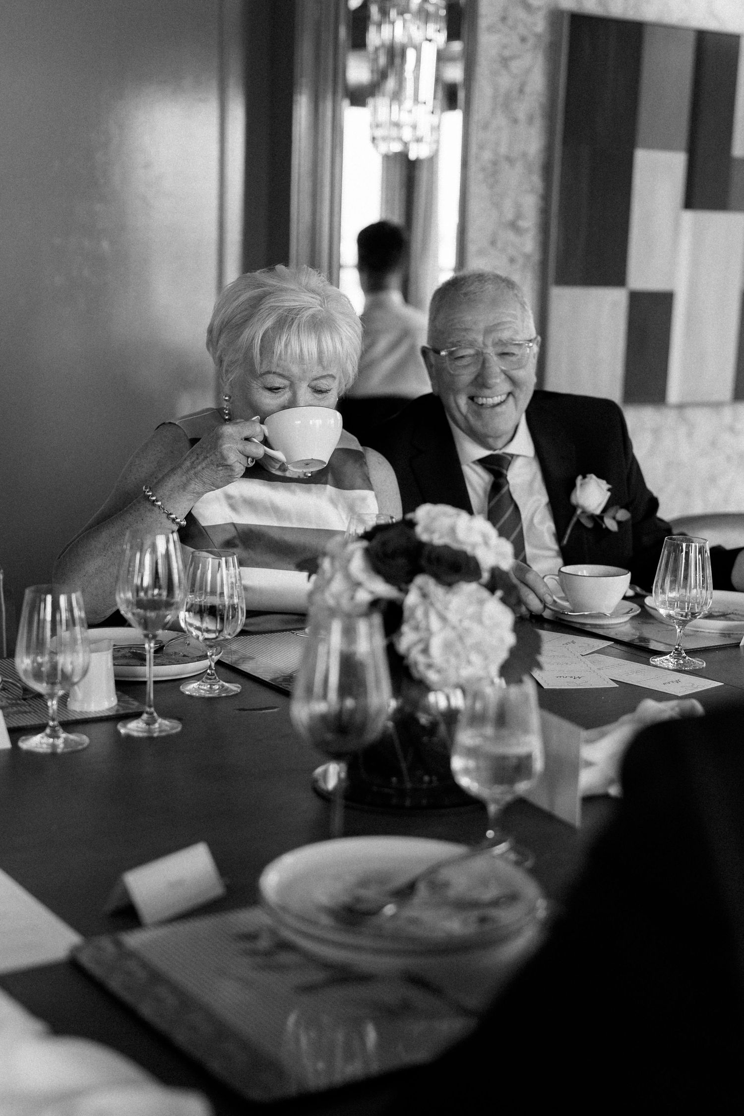 Elderly couple sharing a joyful moment at wedding reception table, woman sipping tea while man smiles warmly.