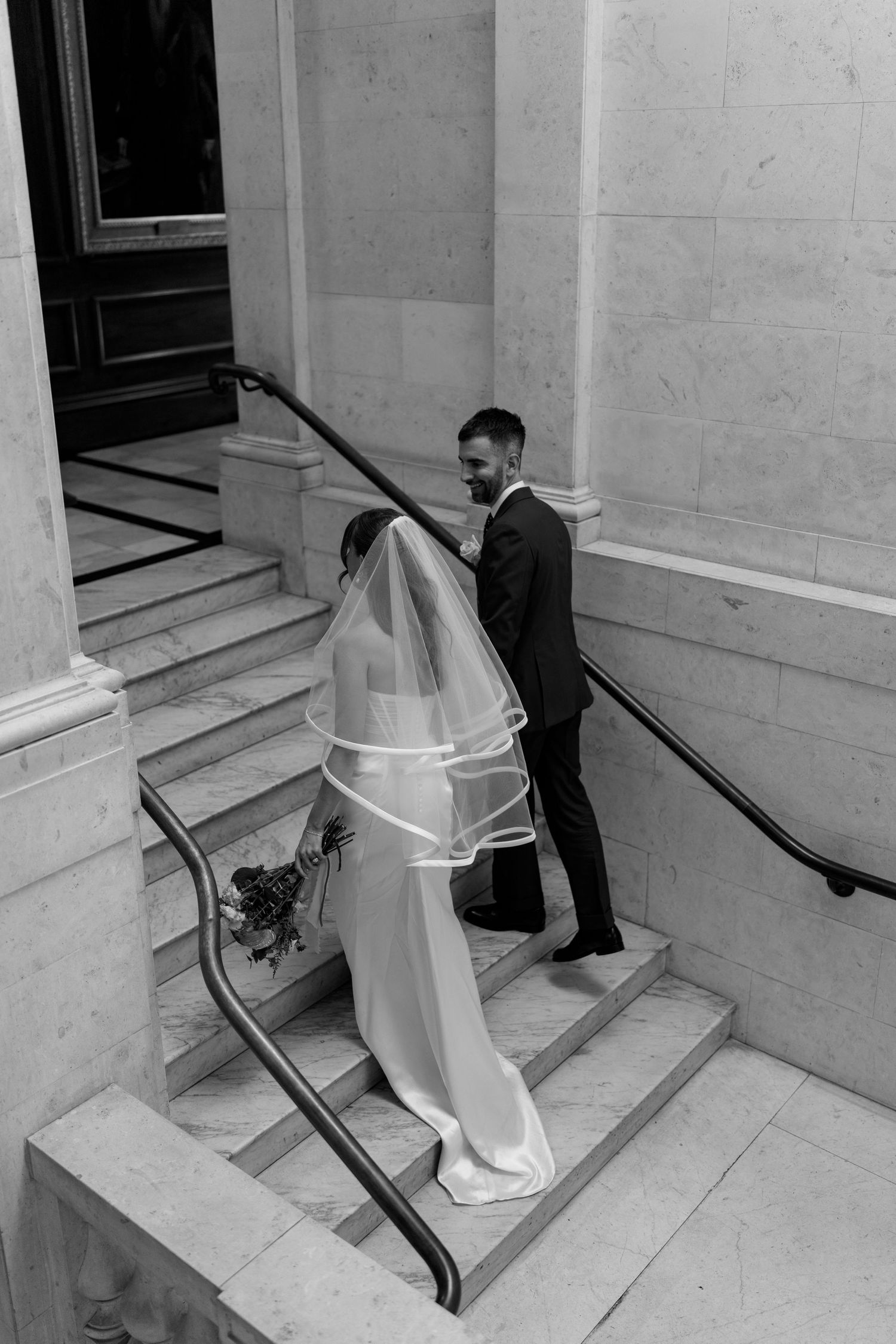 Black and white image of bride and groom walking up elegant marble steps, veil trailing behind.