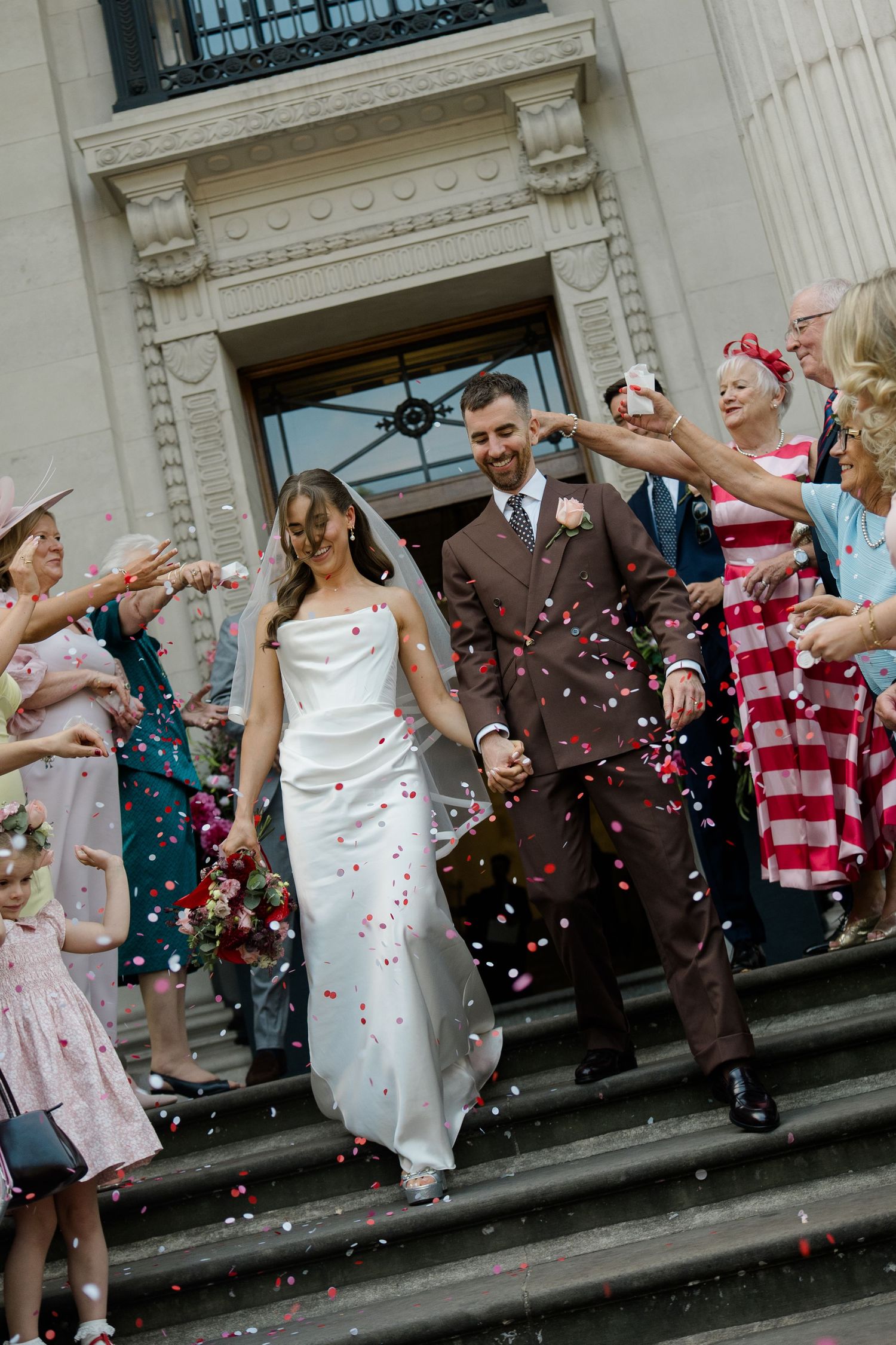 Bride and groom exit registry office steps as guests throw colorful confetti in celebration.