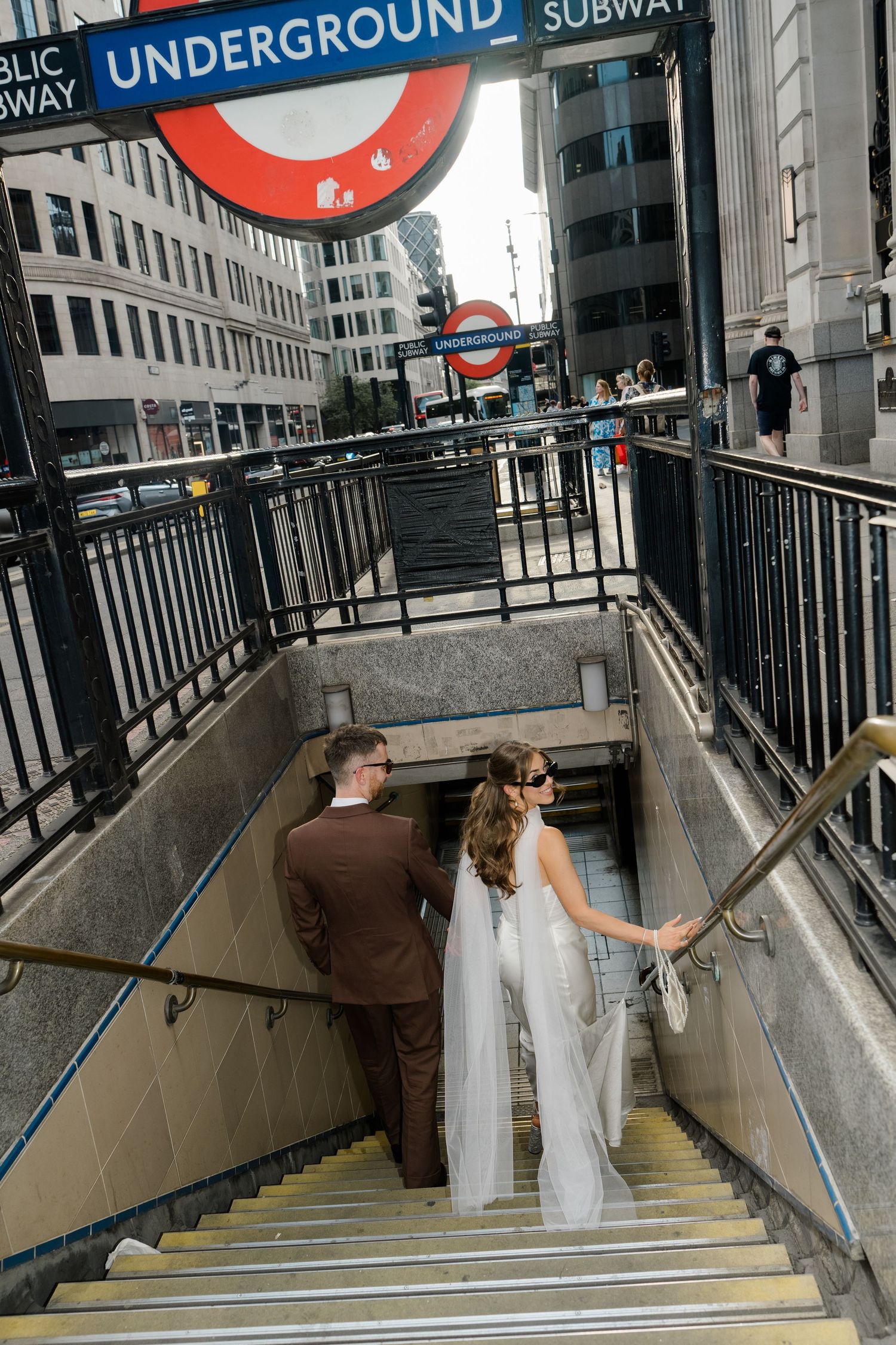 Newlywed couple descending stairs at a London Underground subway station in wedding attire.