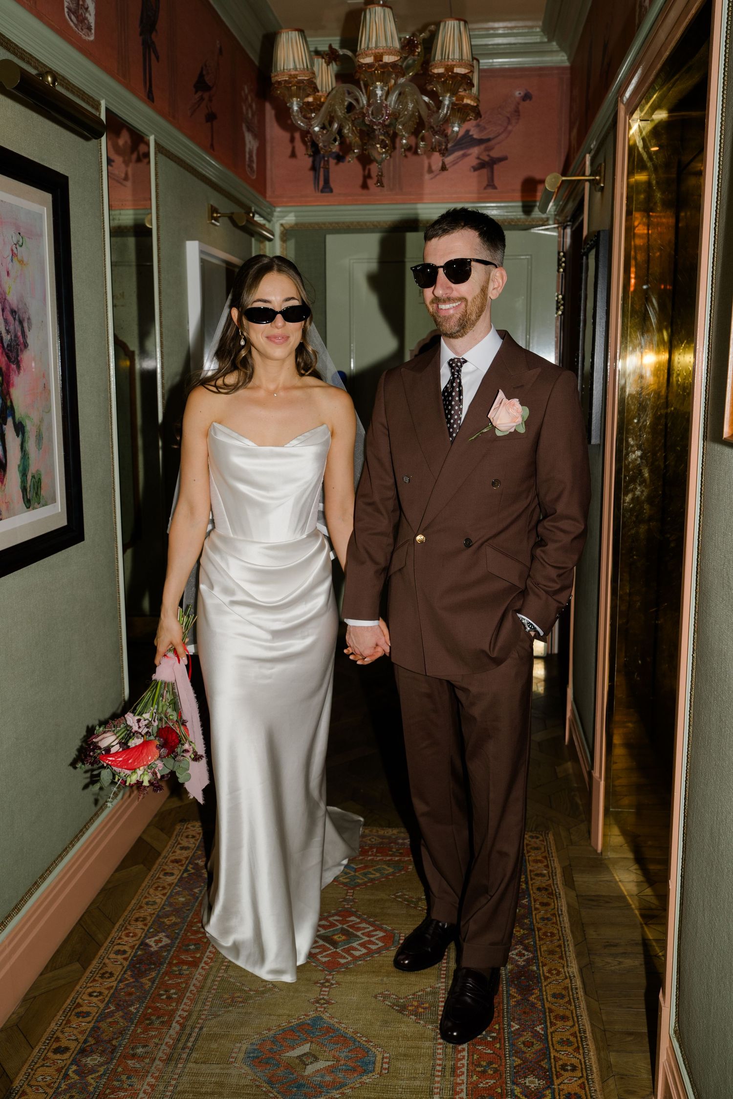 Stylish bride and groom in sunglasses hold hands walking through ornate hotel corridor.