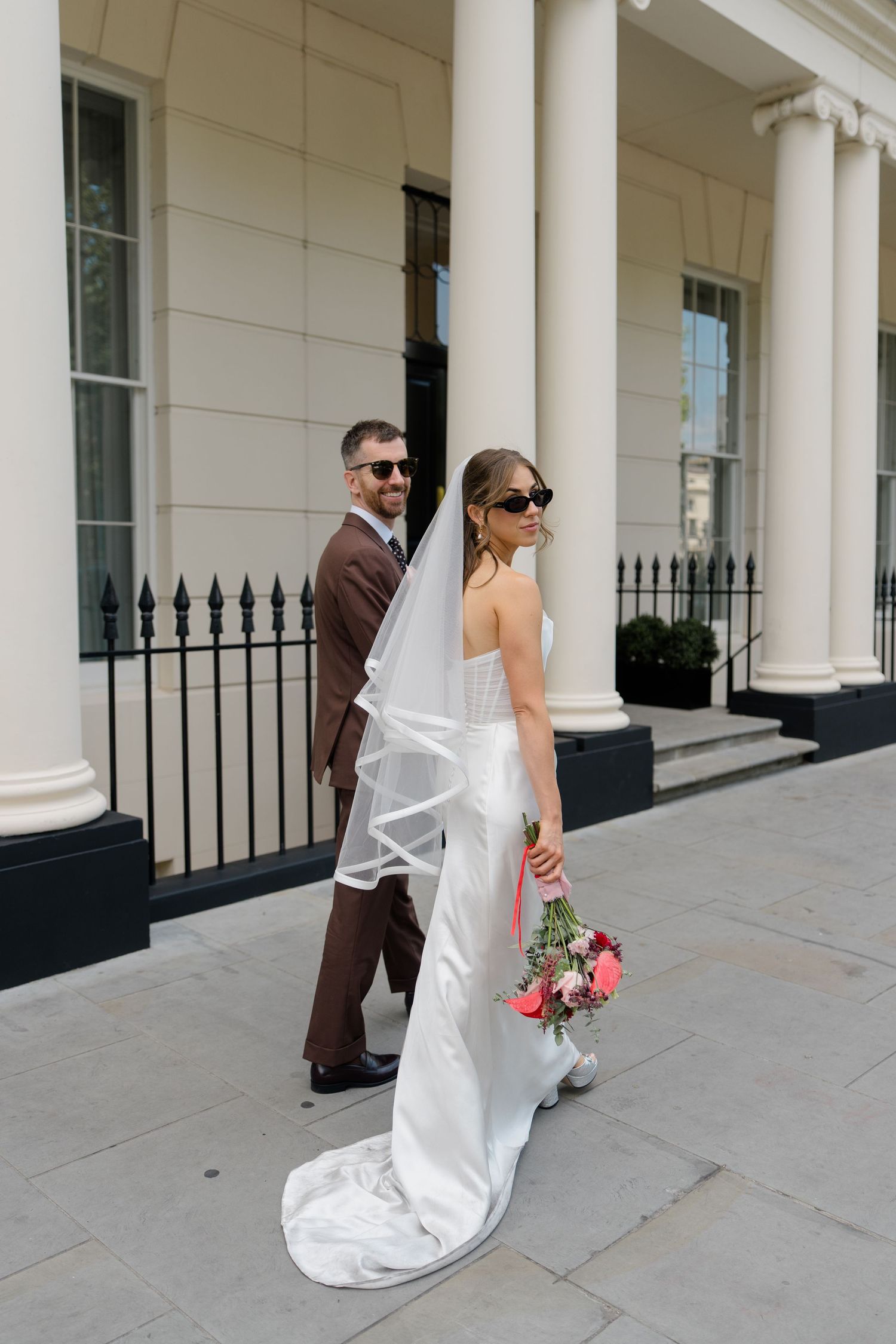 Bride in white gown with veil and sunglasses poses with groom in brown suit outside white Georgian-style building.