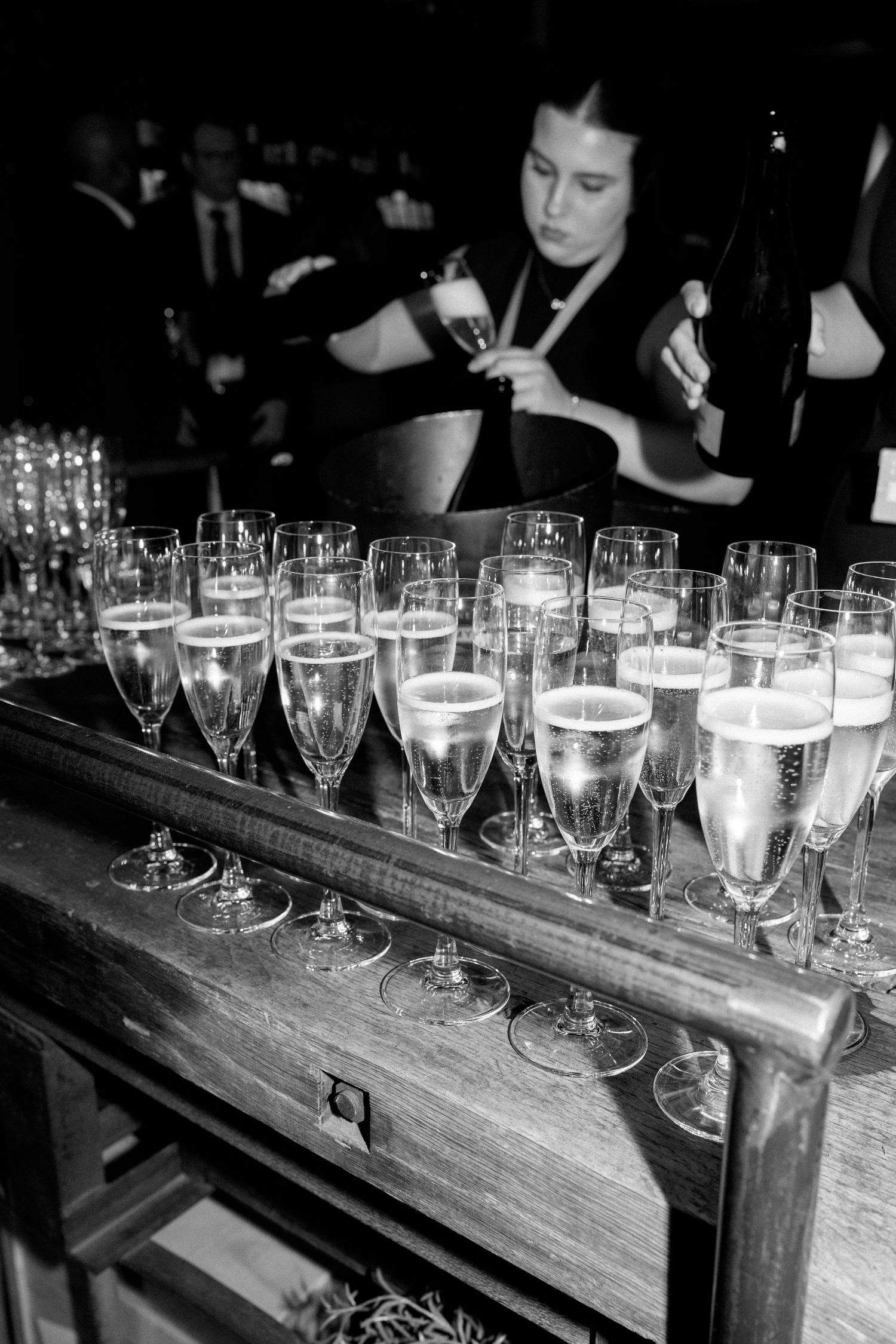Black and white photo of champagne flutes arranged on wooden bar cart at wedding reception.