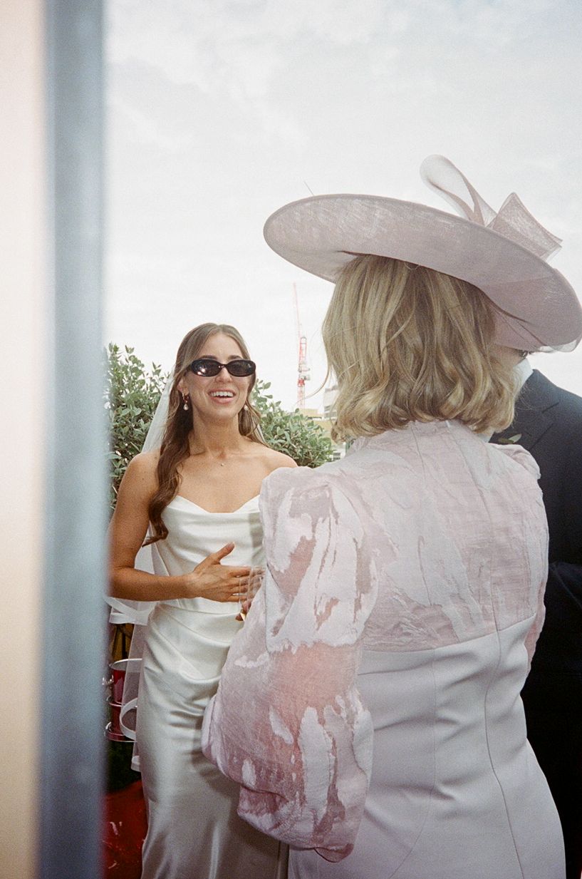 Smiling bride in white dress and sunglasses greeting guest in pink hat and fascinator.