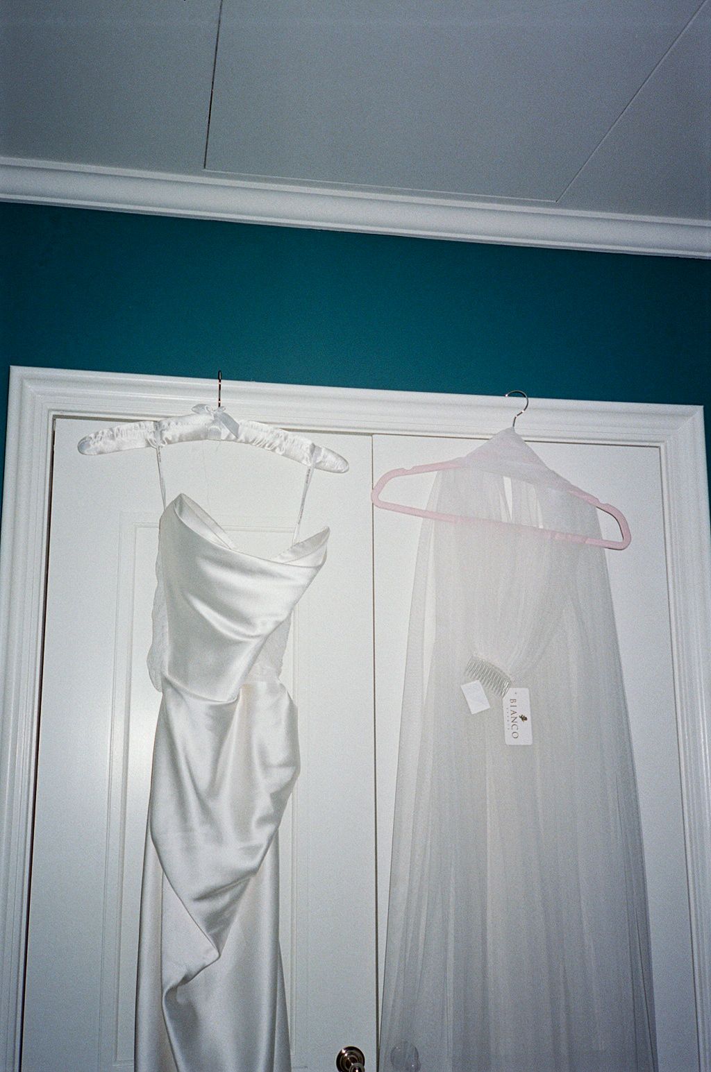 Two white dresses hanging on a closet door, one satin strapless gown and one sheer veil on pink hanger.