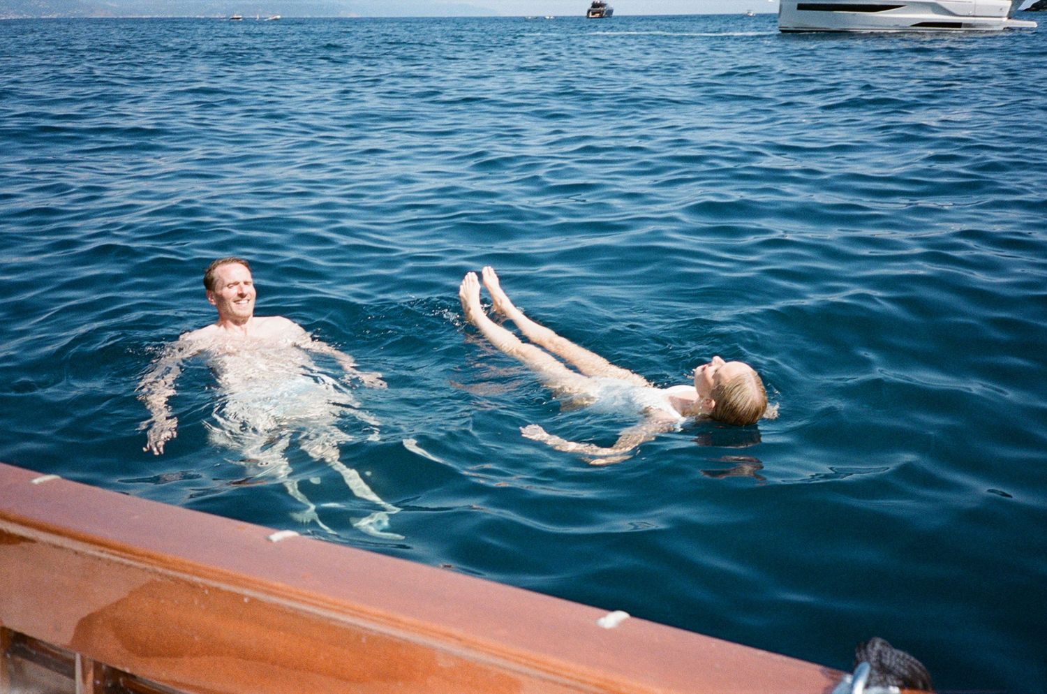 Couple swimming together in deep blue Mediterranean sea beside wooden luxury boat on sunny day.