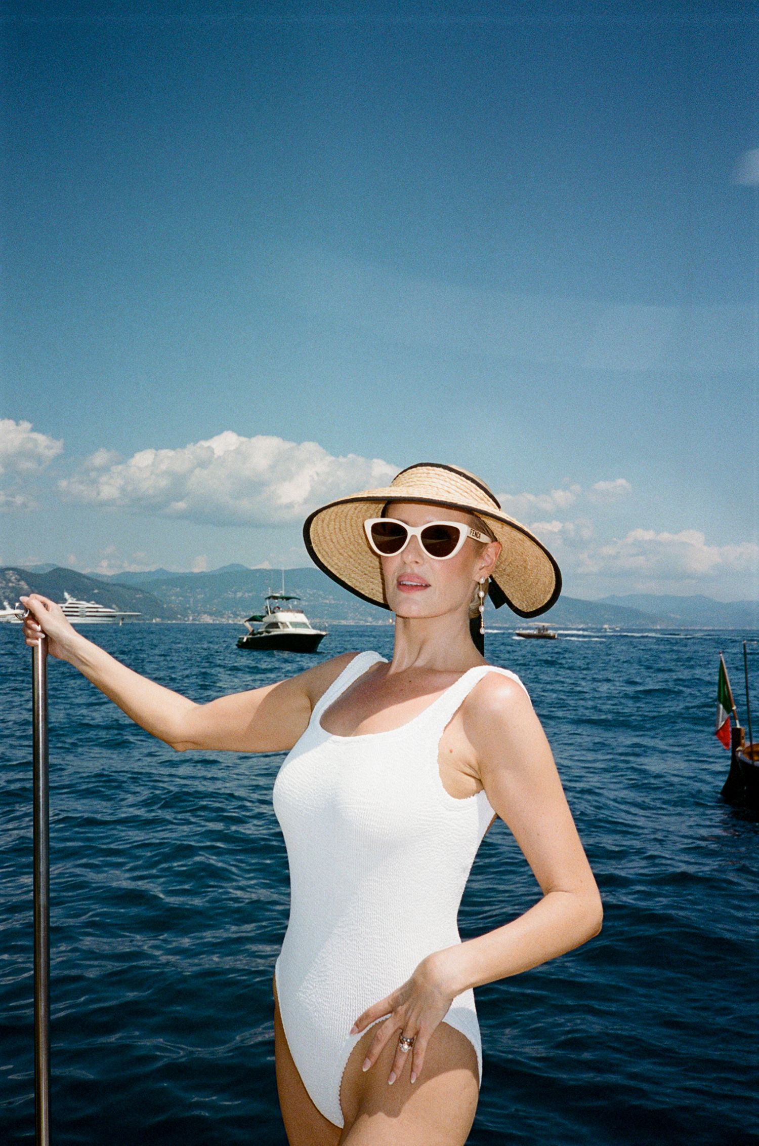 Woman in white swimsuit and wide-brim straw hat with cat-eye sunglasses posing on boat on blue sea.