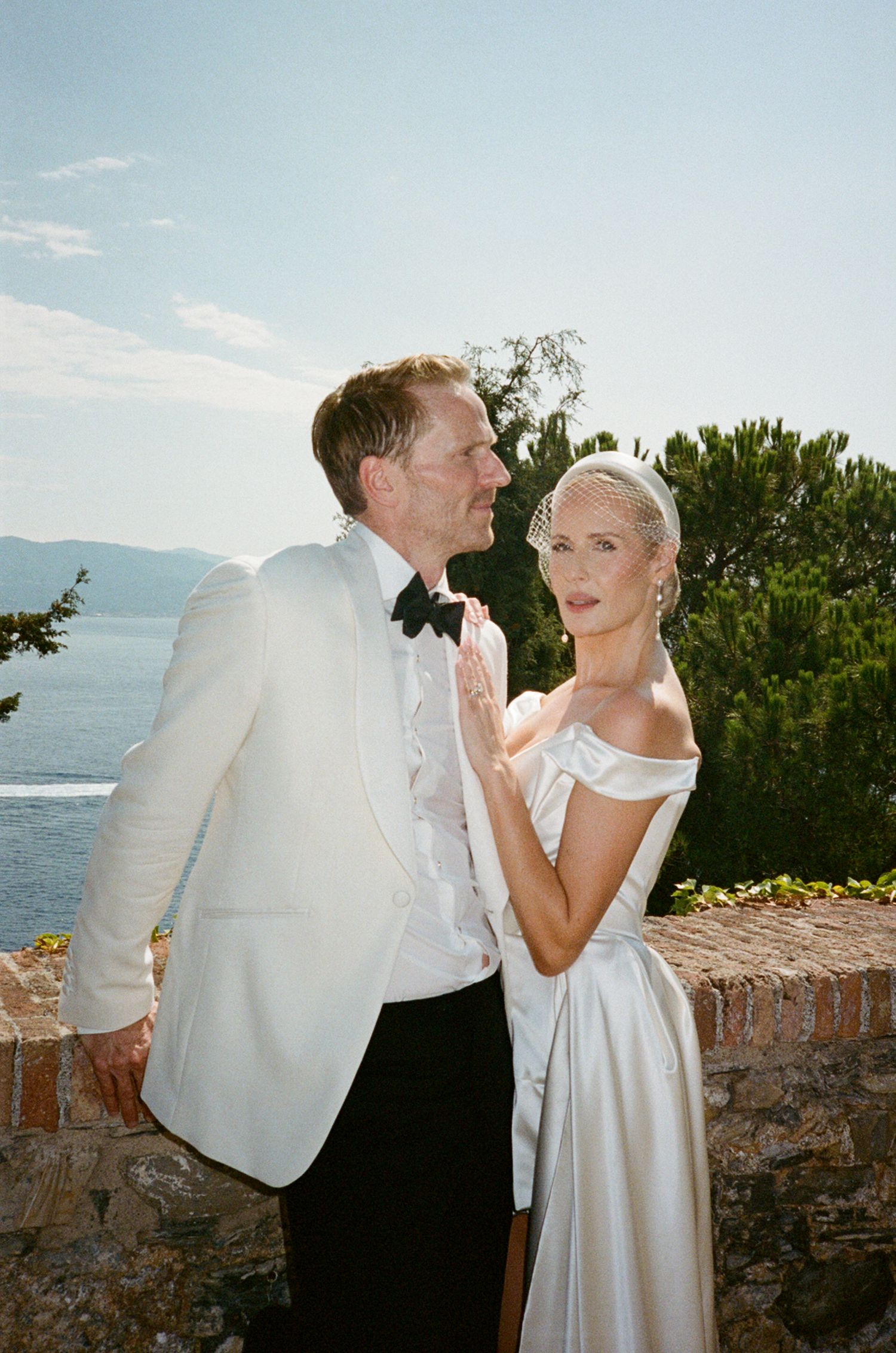 Bride and groom in white wedding attire embracing on terrace overlooking blue Mediterranean sea.
