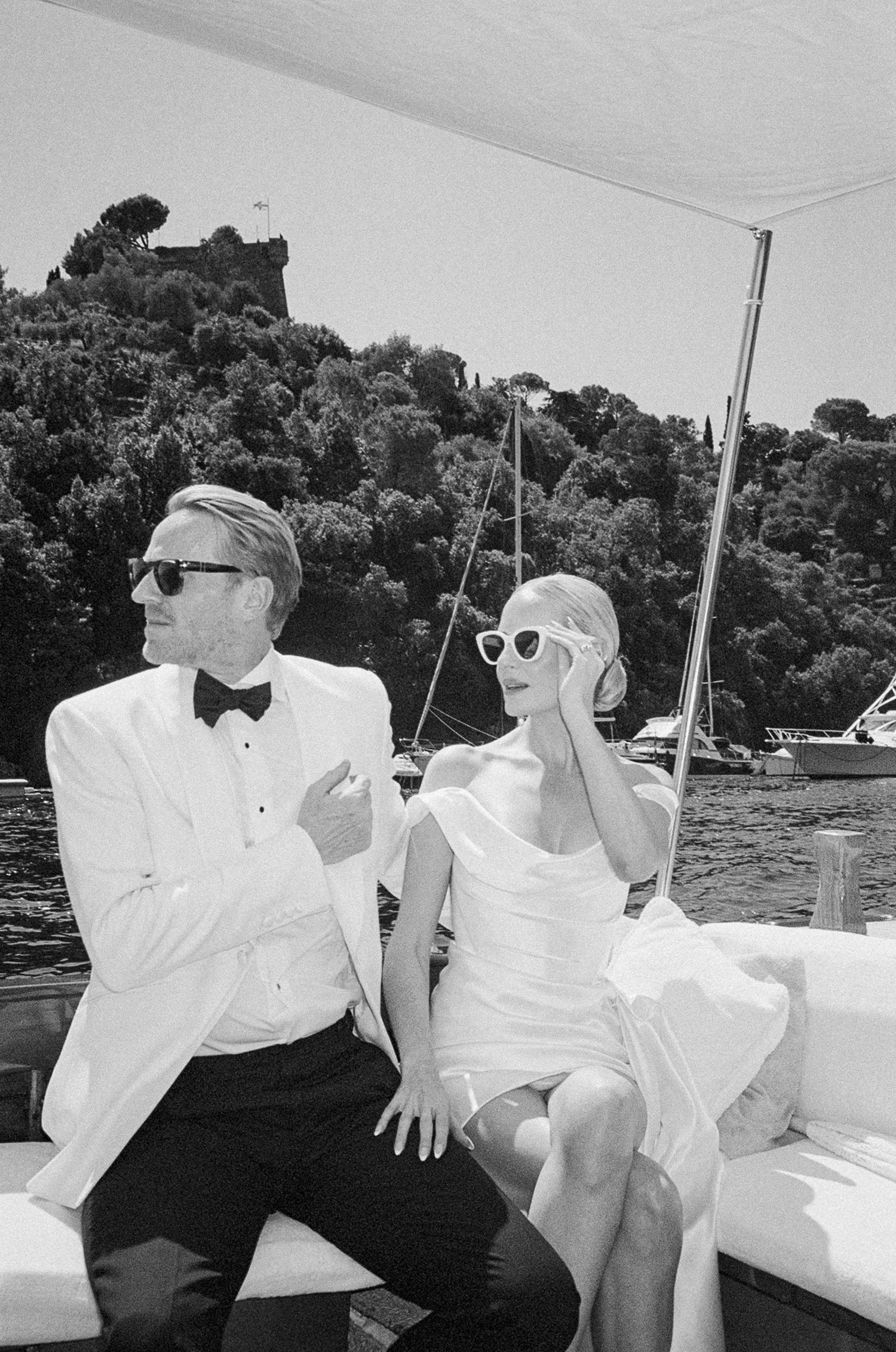 Black and white photo of bride and groom in sunglasses relaxing on a boat near a lush Italian coastline.