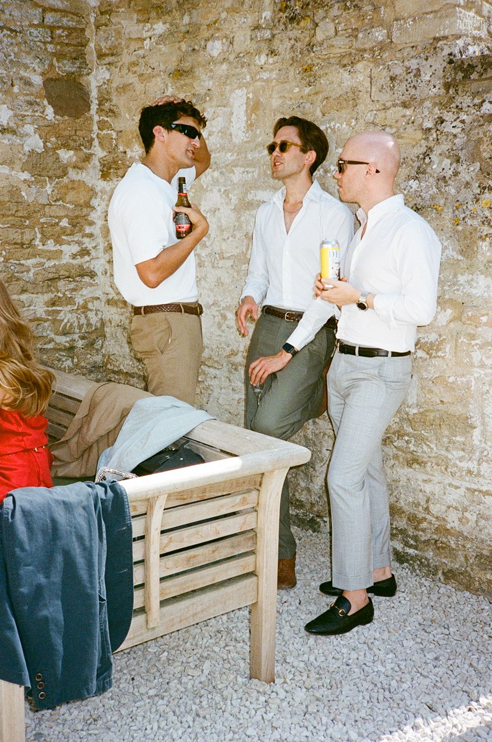 Three men in white shirts and sunglasses socializing outdoors with drinks near a rustic stone wall.