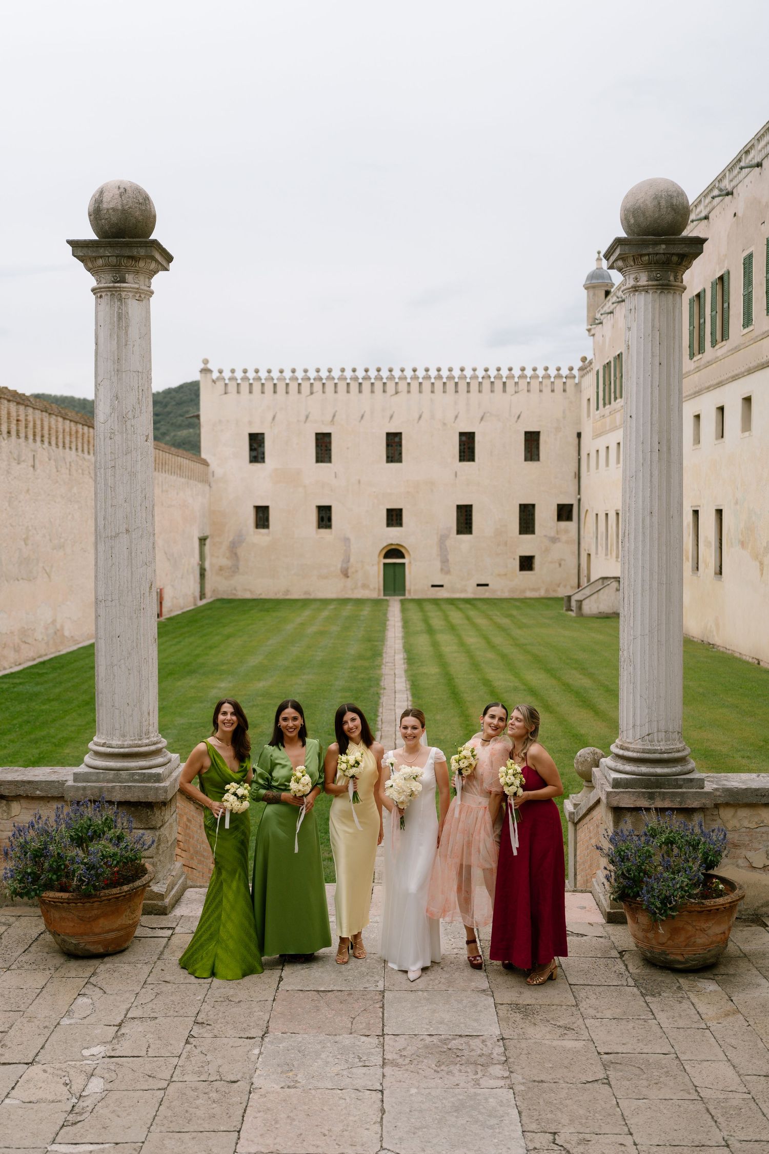 Wedding party in colorful dresses poses between classical columns in a historic courtyard with manicured lawn.