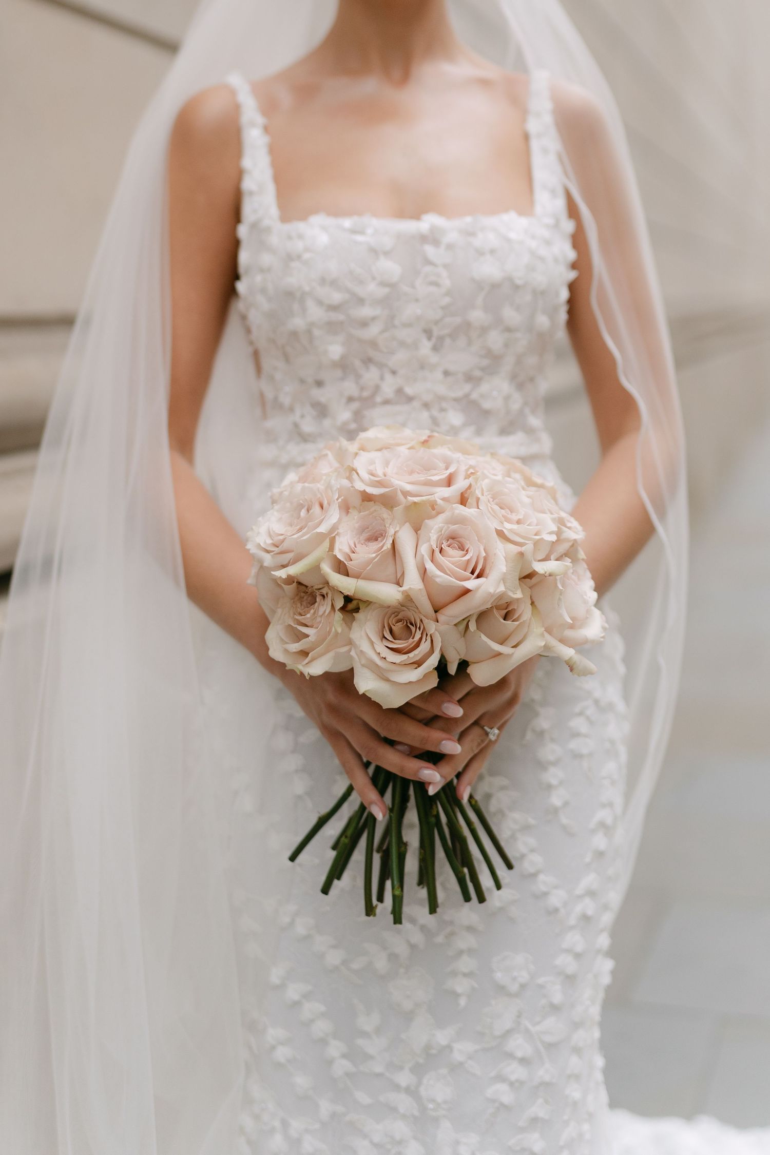 Bride holding delicate bouquet of pale pink roses with lace dress detail.