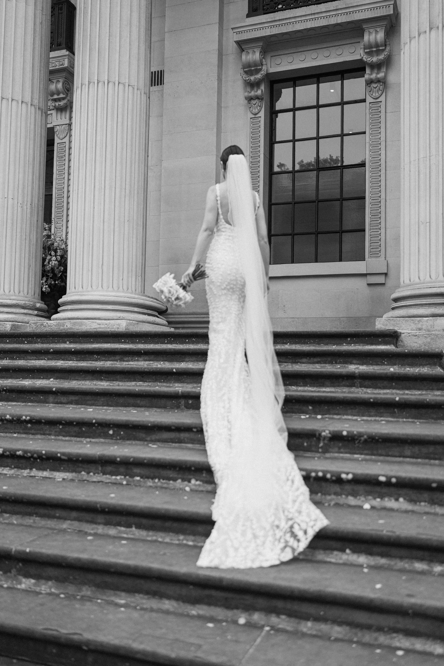 A bride in a fitted lace gown ascends marble steps between ornate white columns.