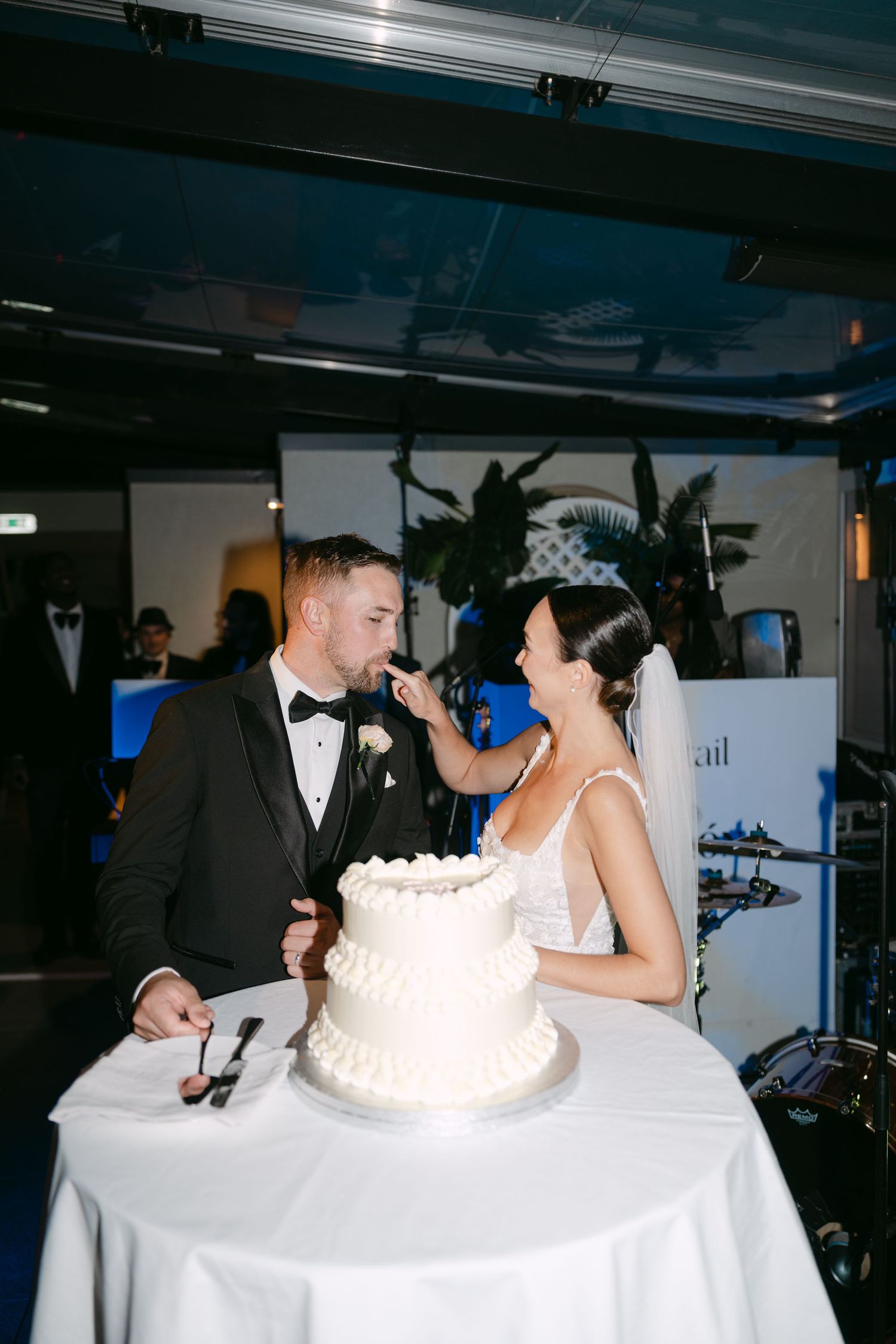 Playful cake-cutting moment at a wedding reception with decorative palm plants in background.