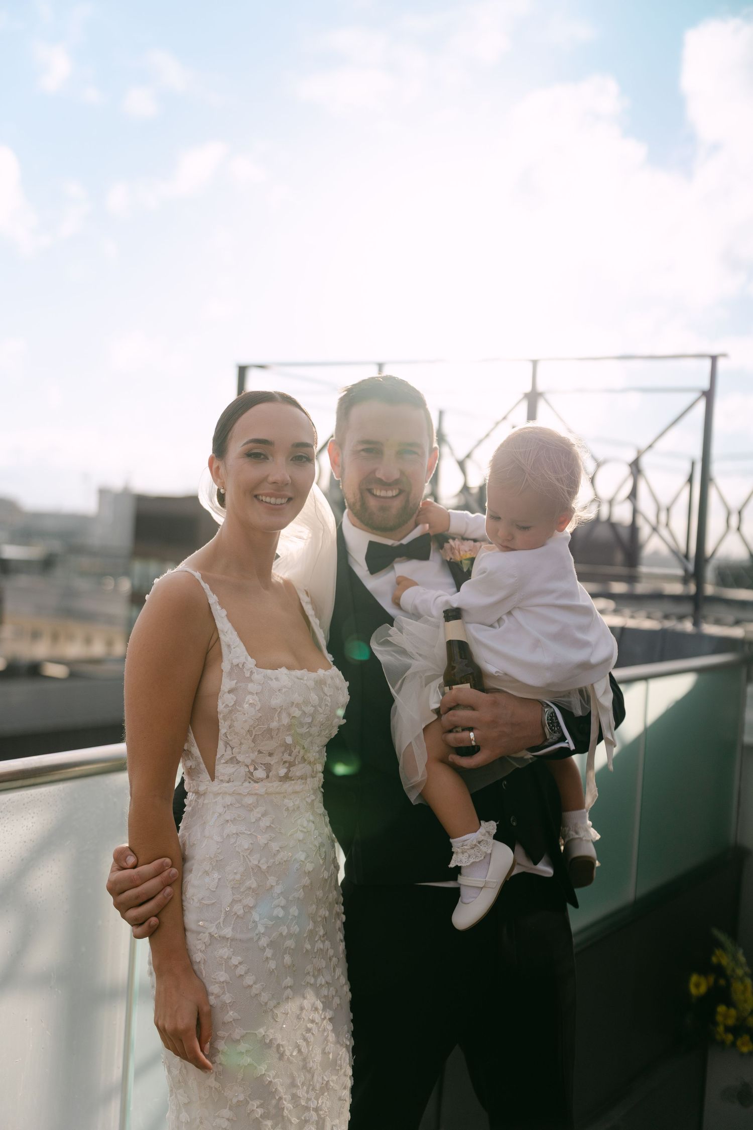 A wedding couple poses with a baby on a rooftop terrace against an urban backdrop.